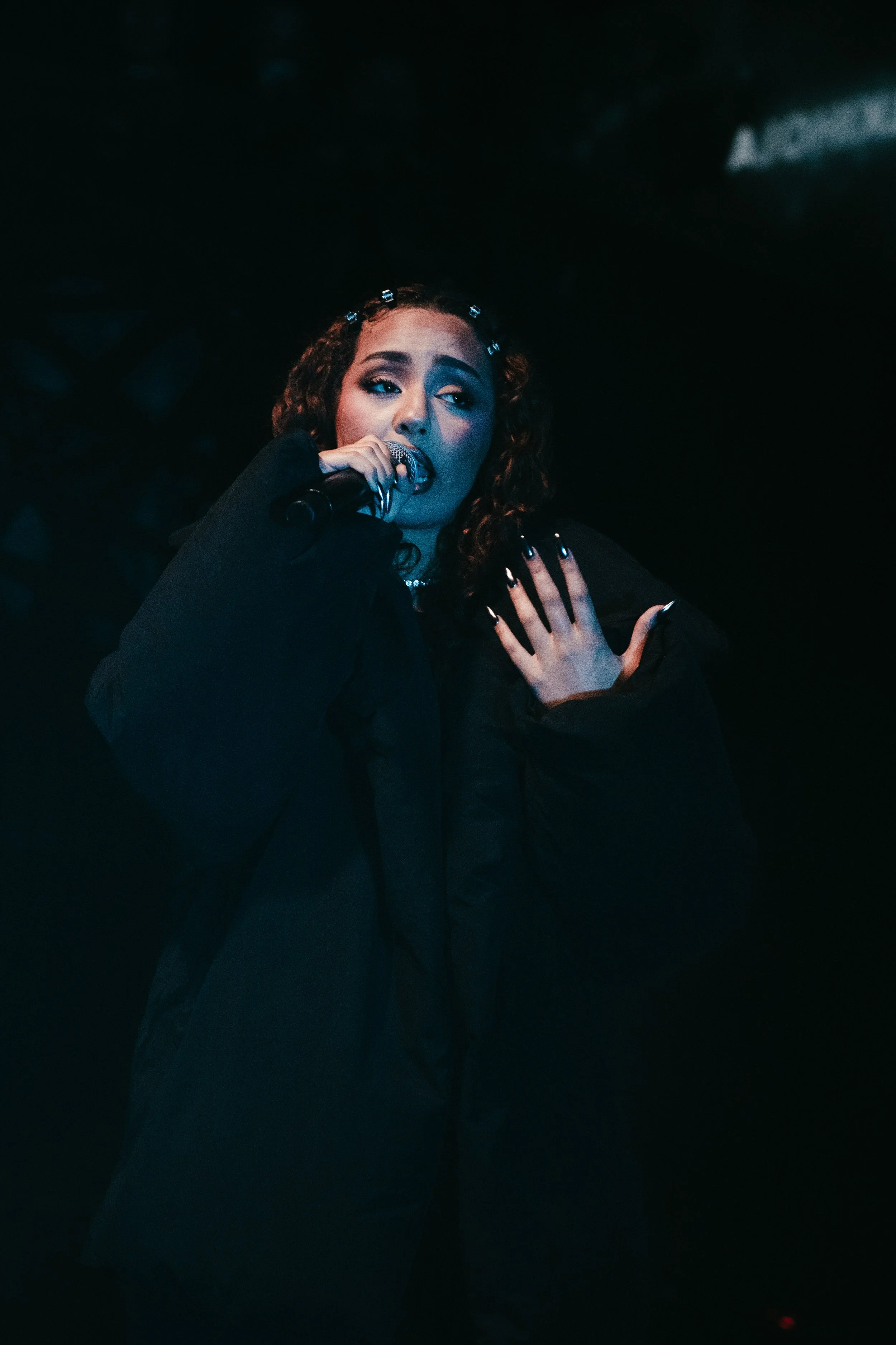 Une jeune femme chante dans un microphone lors d'une performance sur scène, portant un vêtement noir, avec des cheveux bouclés et un maquillage marqué.