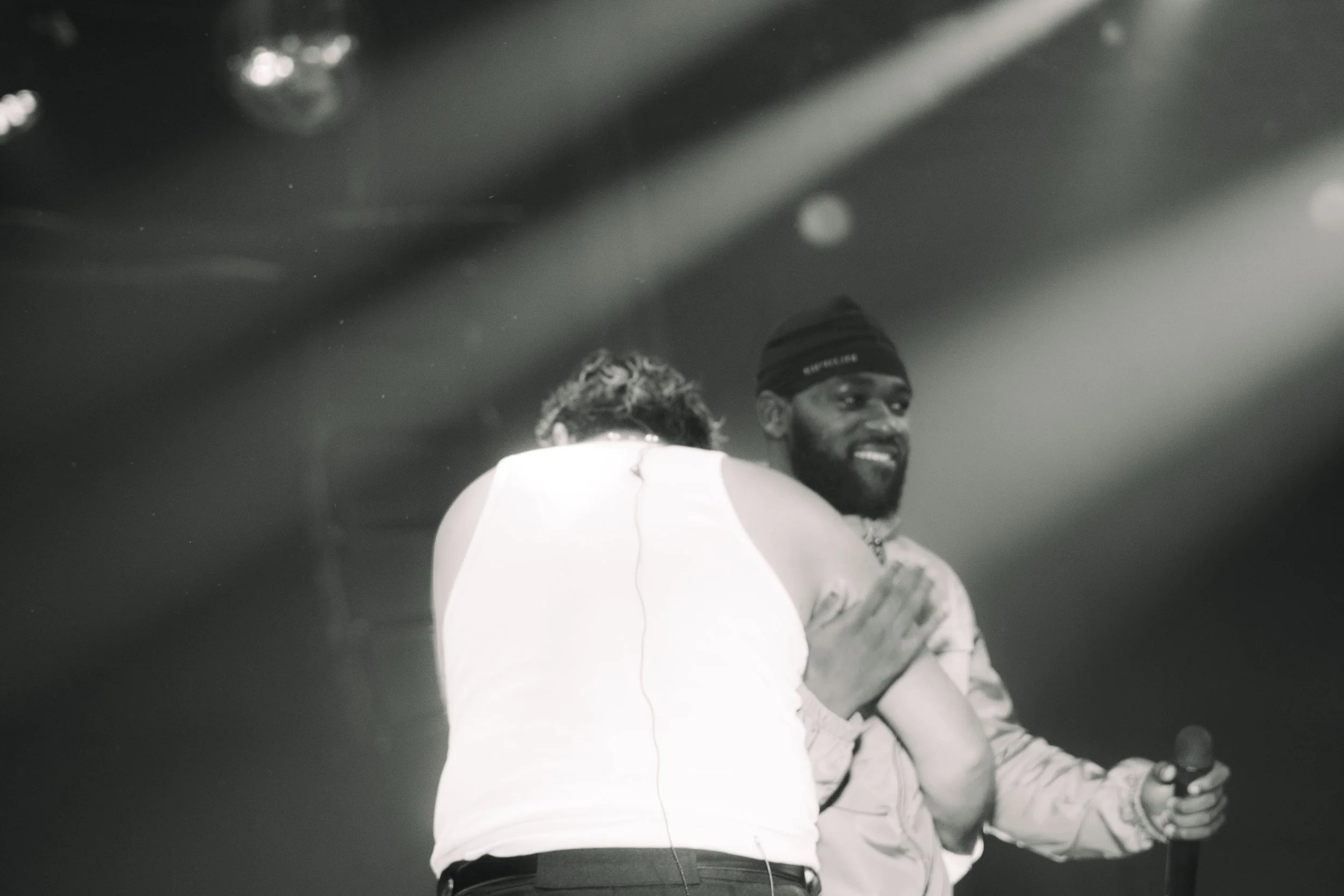 Deux hommes sur scène, l'un de dos et l'autre de face, en train de se sourire. L'homme de face tient un micro et porte un bonnet. L'image est en noir et blanc.