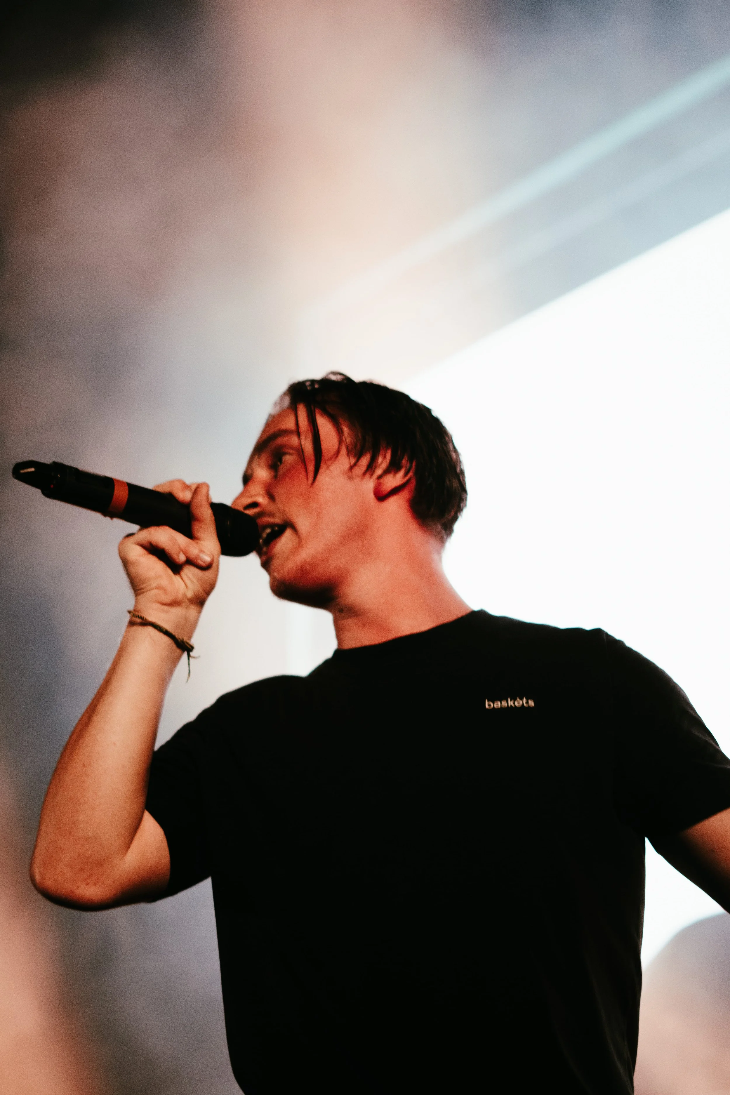Une personne tenant un microphone en chantant ou parlant lors d'une performance sur scène, vêtue d'un t-shirt noir.