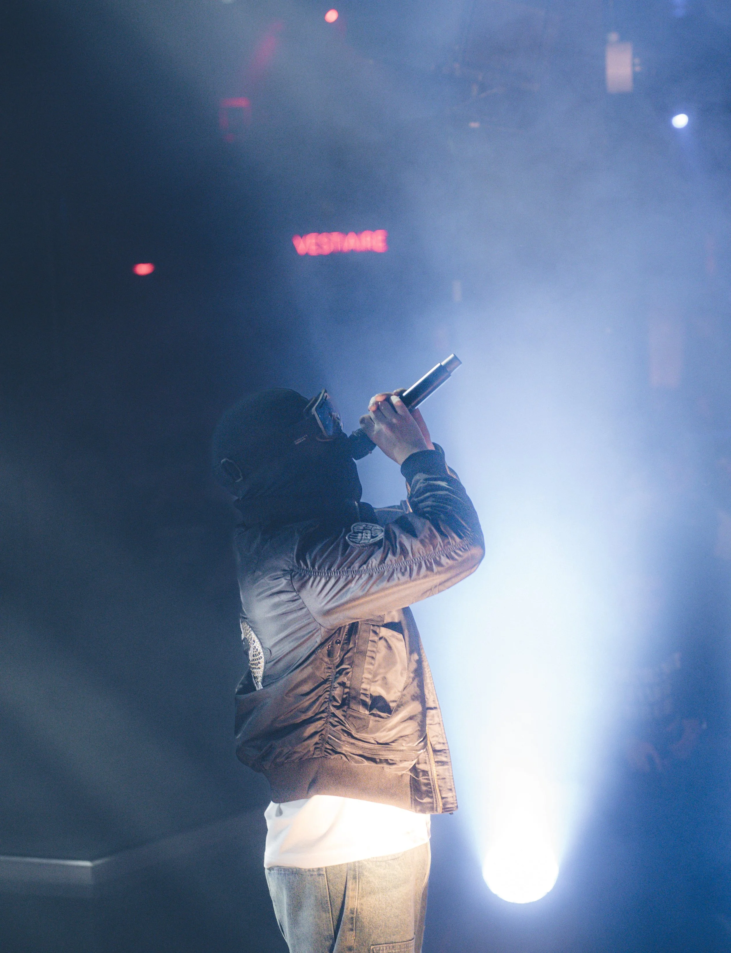 Personne portant un masque et des lunettes de protection, avec une veste en satin marron clair, tenant un microphone sur scène avec un éclairage de scène brillante.