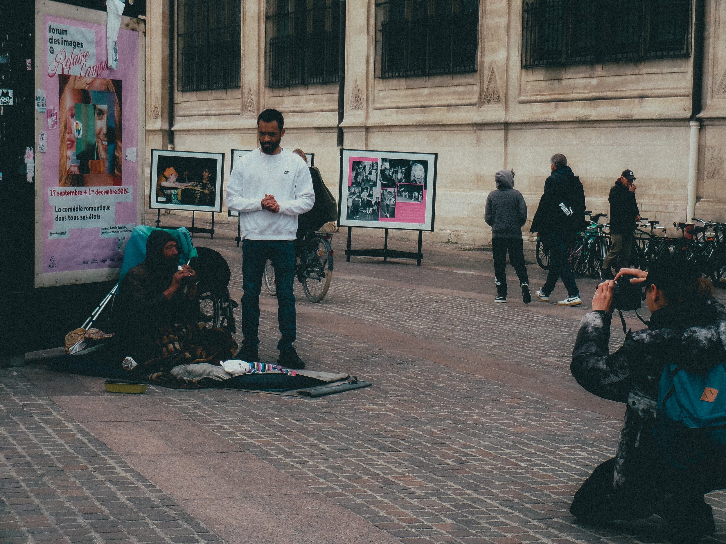 Une personne sans-abri assise sur un tapis au sol, avec un homme debout devant elle, dans une rue pavée, à côté d'une affiche de spectacle. Un autre homme est photographiant, deux autres personnes marchent en arrière-plan, et plusieurs vélos sont sta