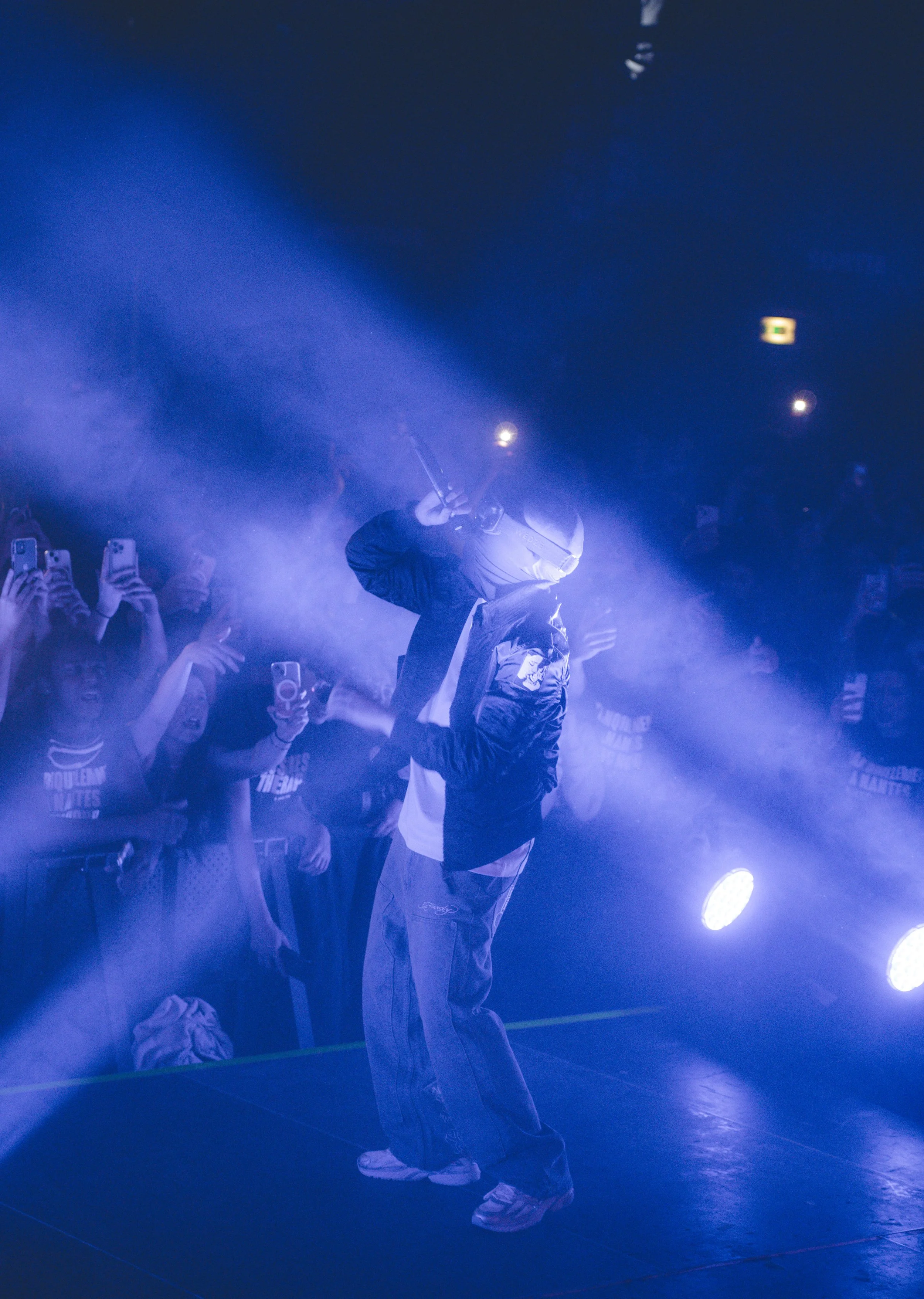 Une personne portant un casque de moto et une veste sombre, tenant un objet dans sa main, se tient sur scène avec un éclairage violet et un brouillard, entourée de spectateurs prenant des photos.