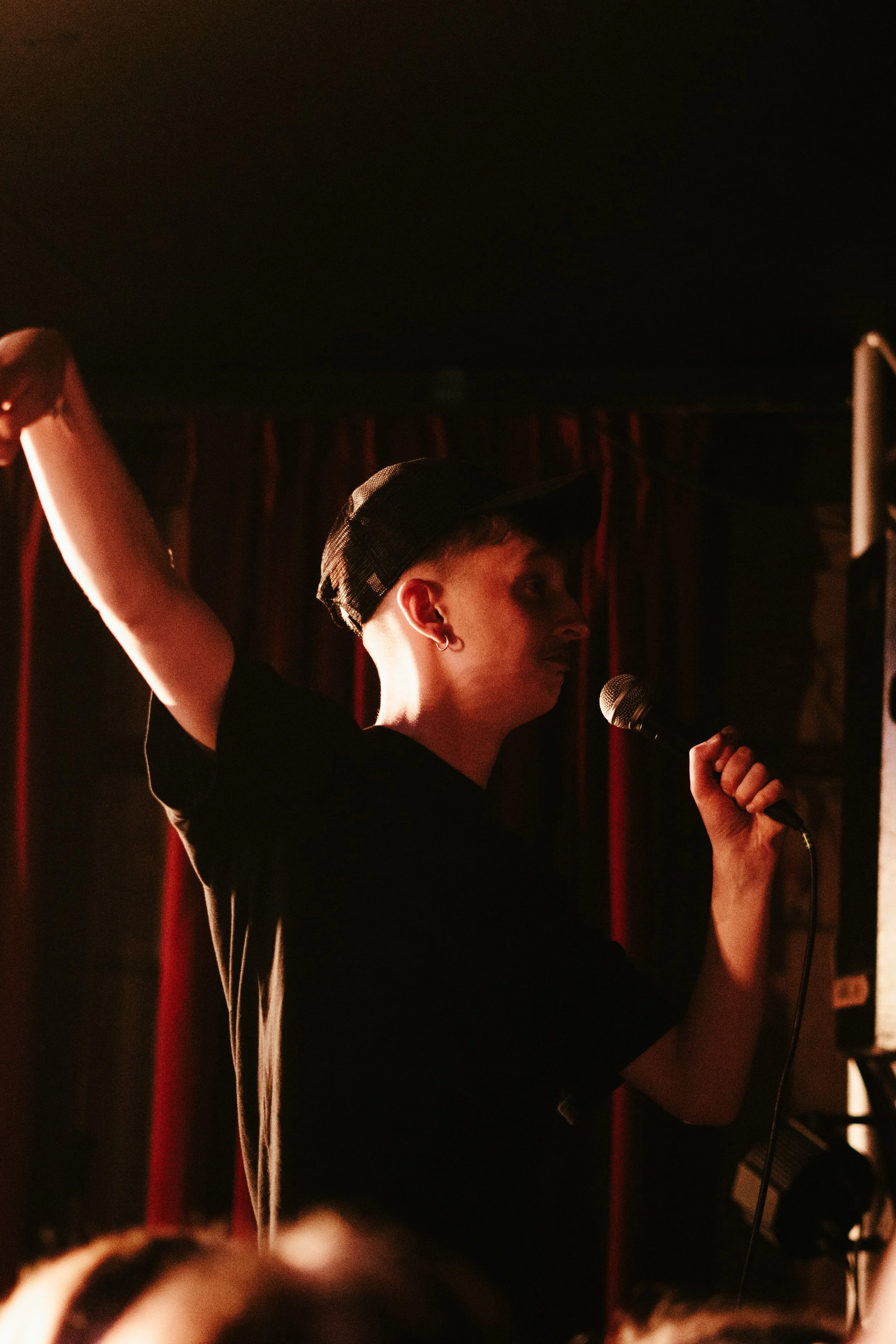 Une personne debout avec un bras levé, tenant un micro, portant une casquette noire, dans un environnement sombre avec des rideaux rouges en arrière-plan.