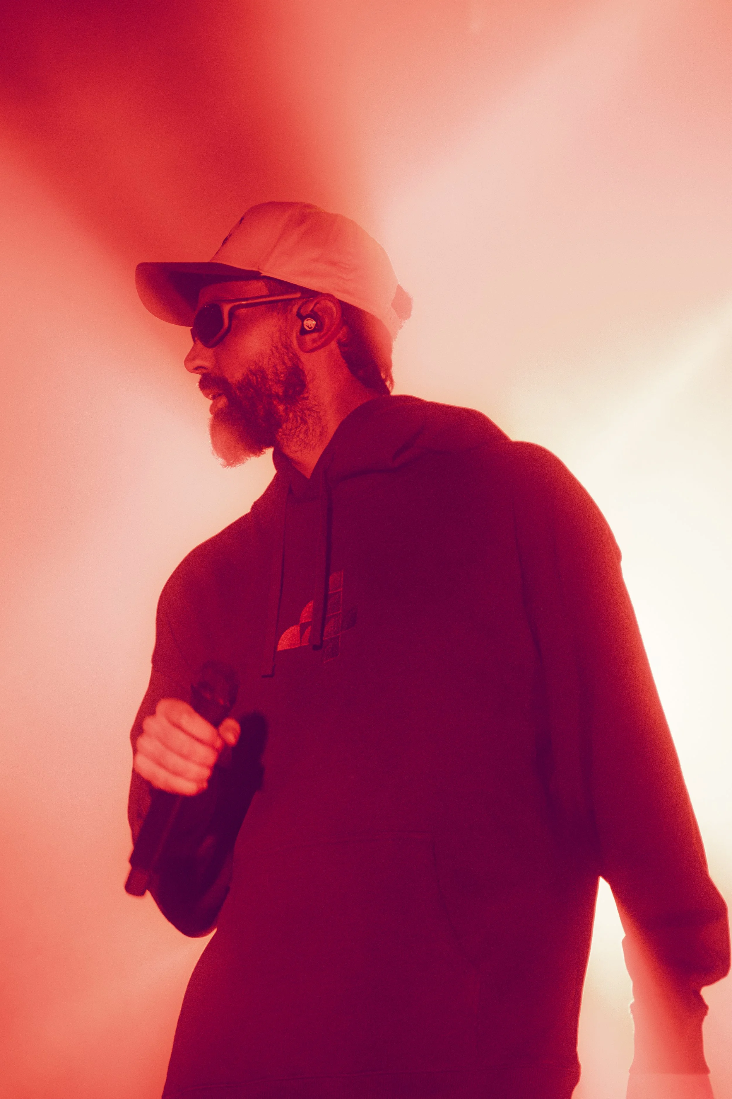 Un homme avec une barbe, portant une casquette, des lunettes de soleil et un sweat à capuche, tenant un microphone, éclairé par une lumière rouge