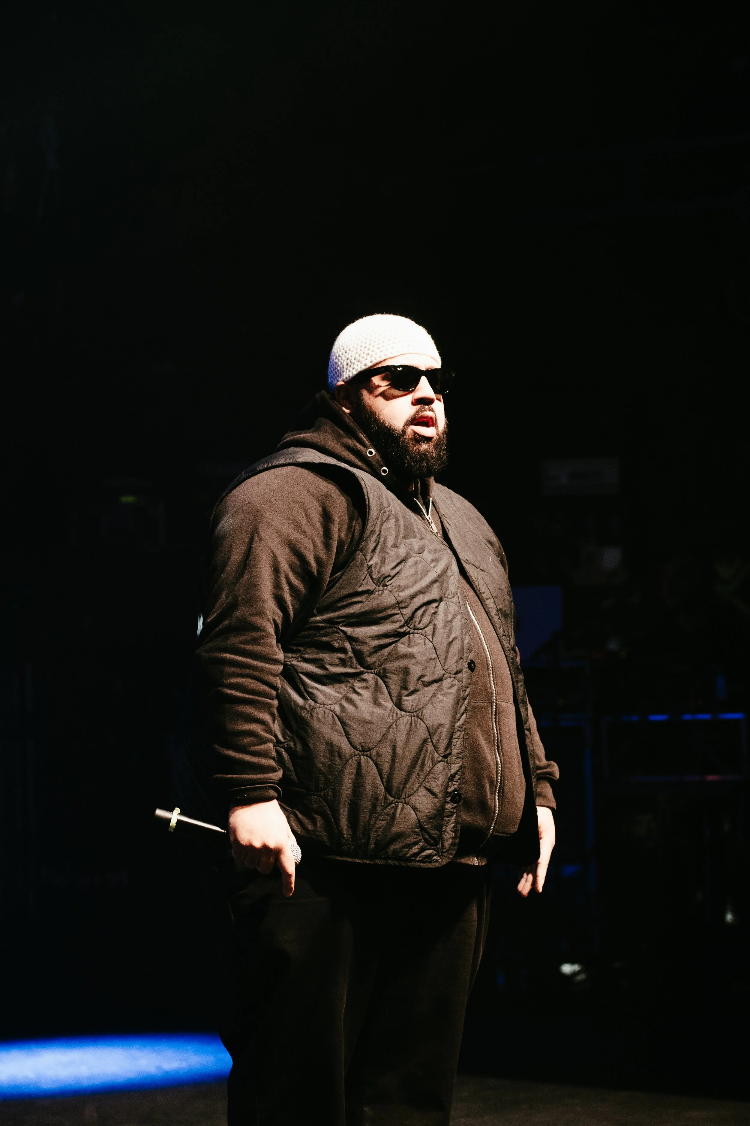 Un homme portant des lunettes de soleil, un bonnet blanc, un haut noir et un manteau brun, se tient dans une scène sombre, probablement lors d'une performance ou d'un spectacle.