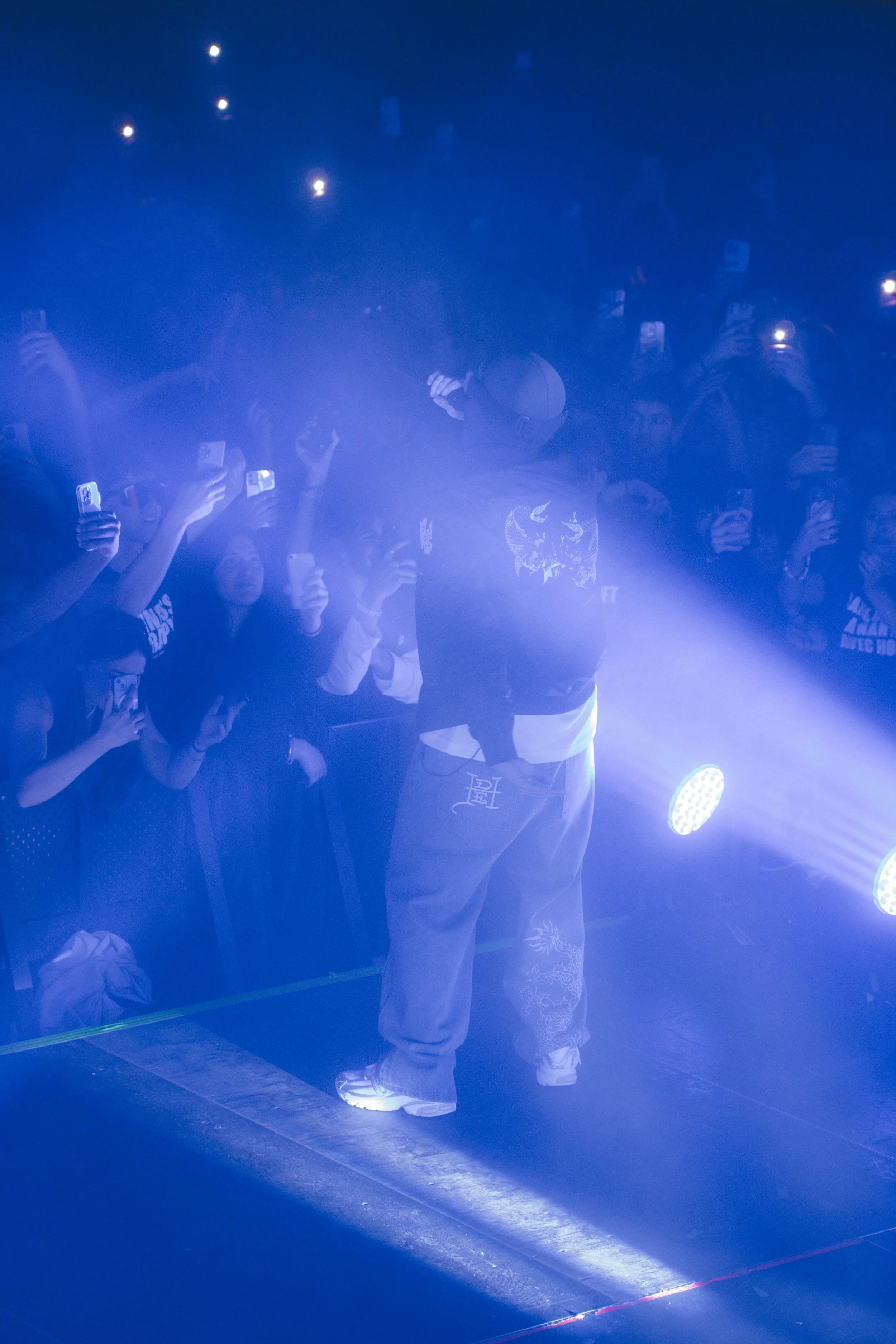 Une personne entre sur scène lors d'un concert ou d'un événement, entourée d'un public qui prend des photos avec leurs téléphones sous un éclairage bleu.