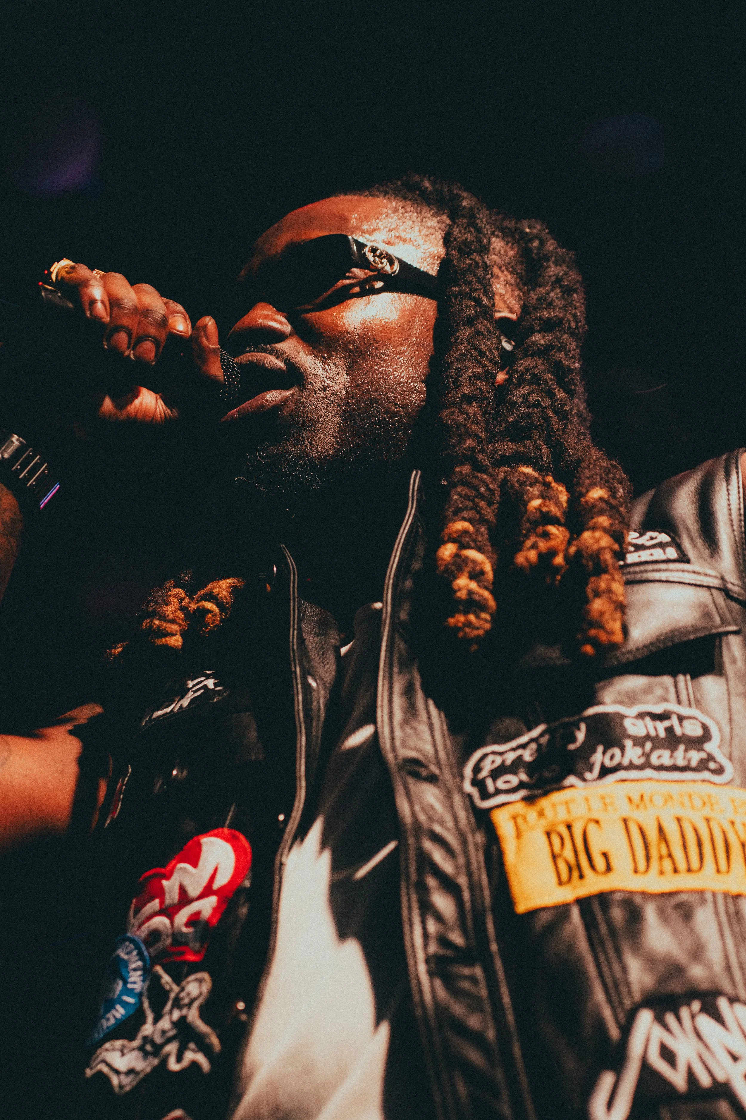 Un homme avec des cheveux en dreadlocks portant des lunettes de soleil et une veste en cuir noire, tenant un microphone près de la bouche, lors d'une performance ou d'un événement.