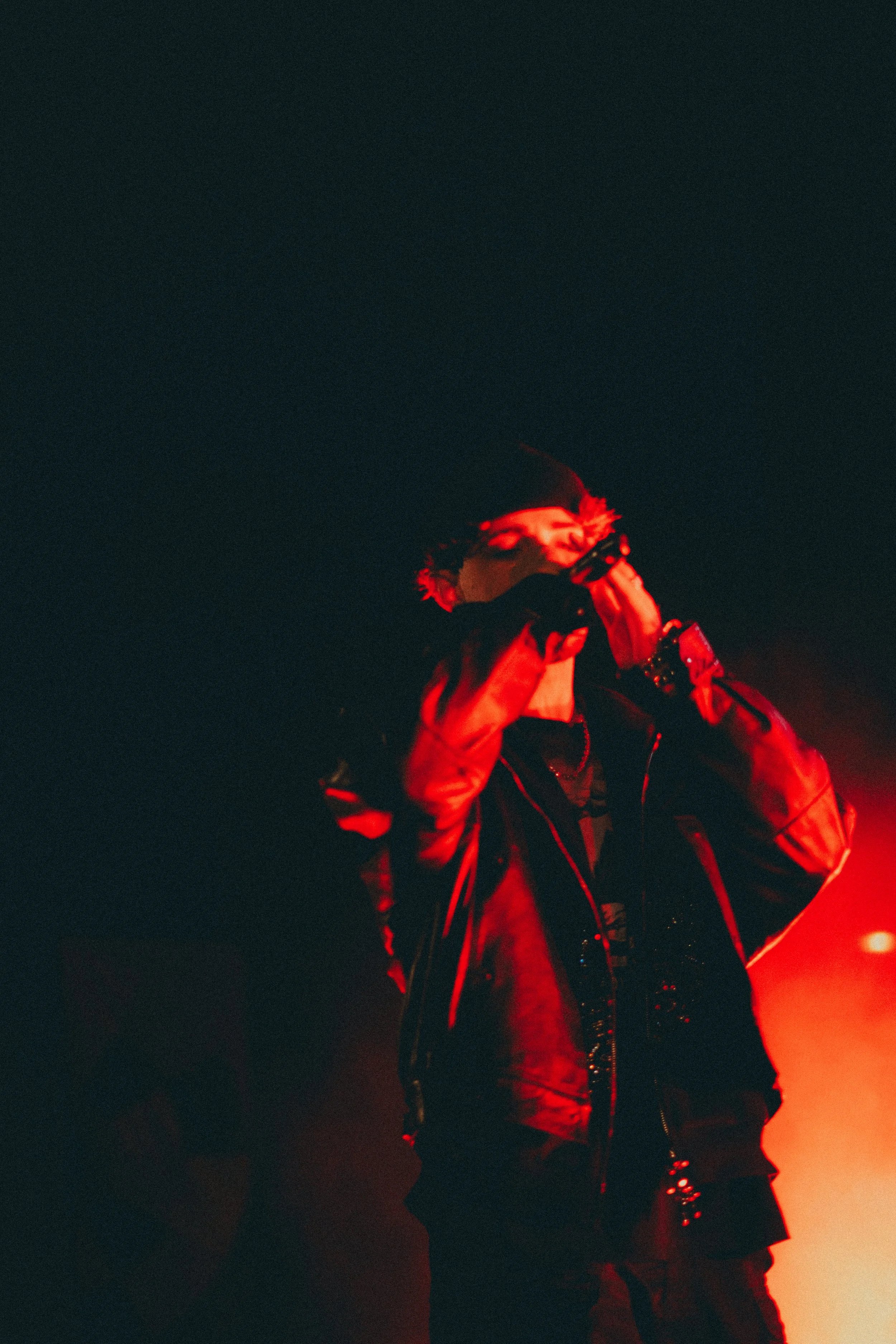 Une personne en vêtements noirs et une veste en cuir, éclairée par une lumière rouge, tenant un micro ou un appareil devant la bouche lors d'une performance ou d'un concert.