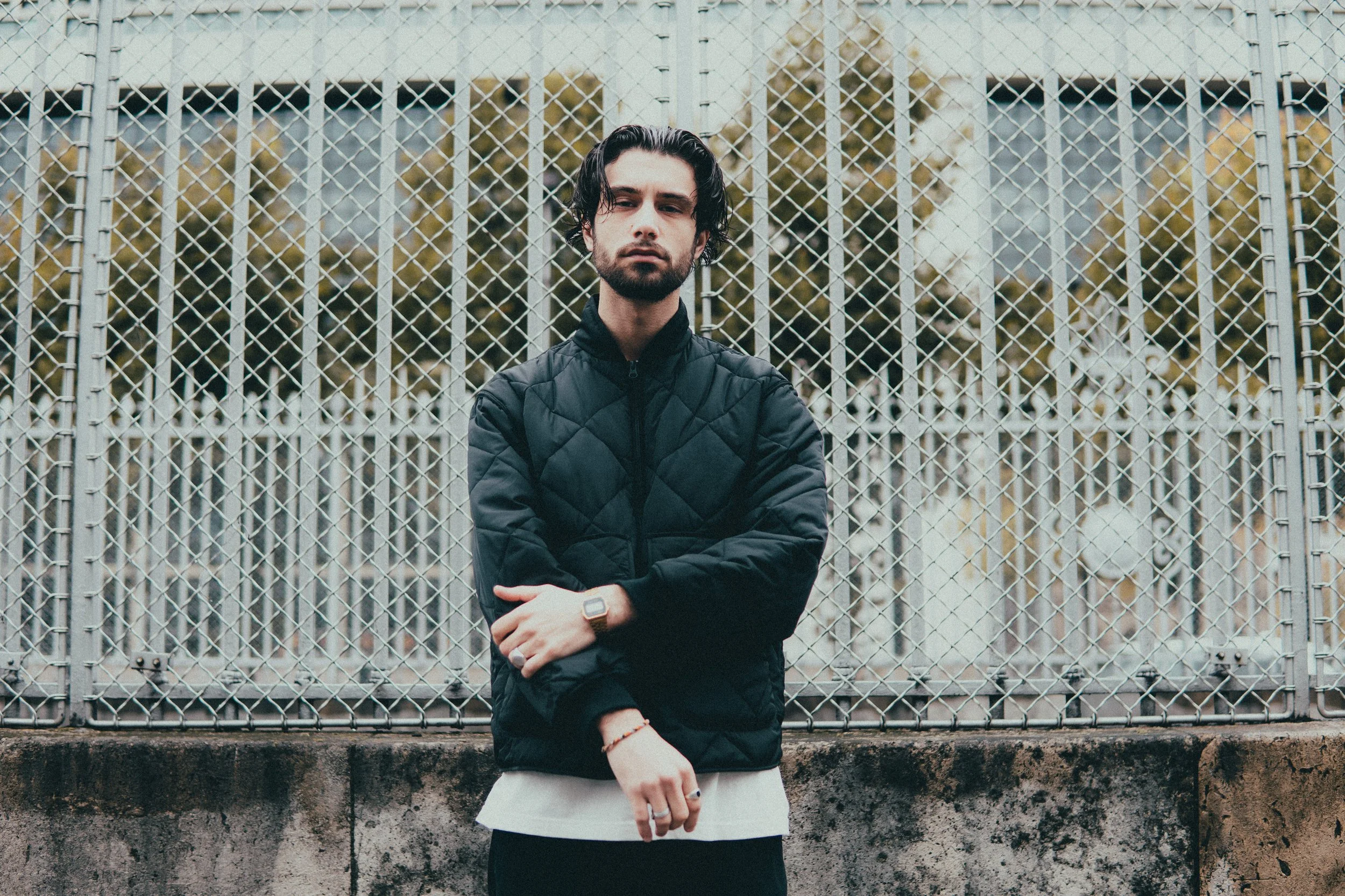 Un homme avec des cheveux noirs, barbe, portant une veste noire, se tient devant une grille en métal, avec des arbres en arrière-plan.