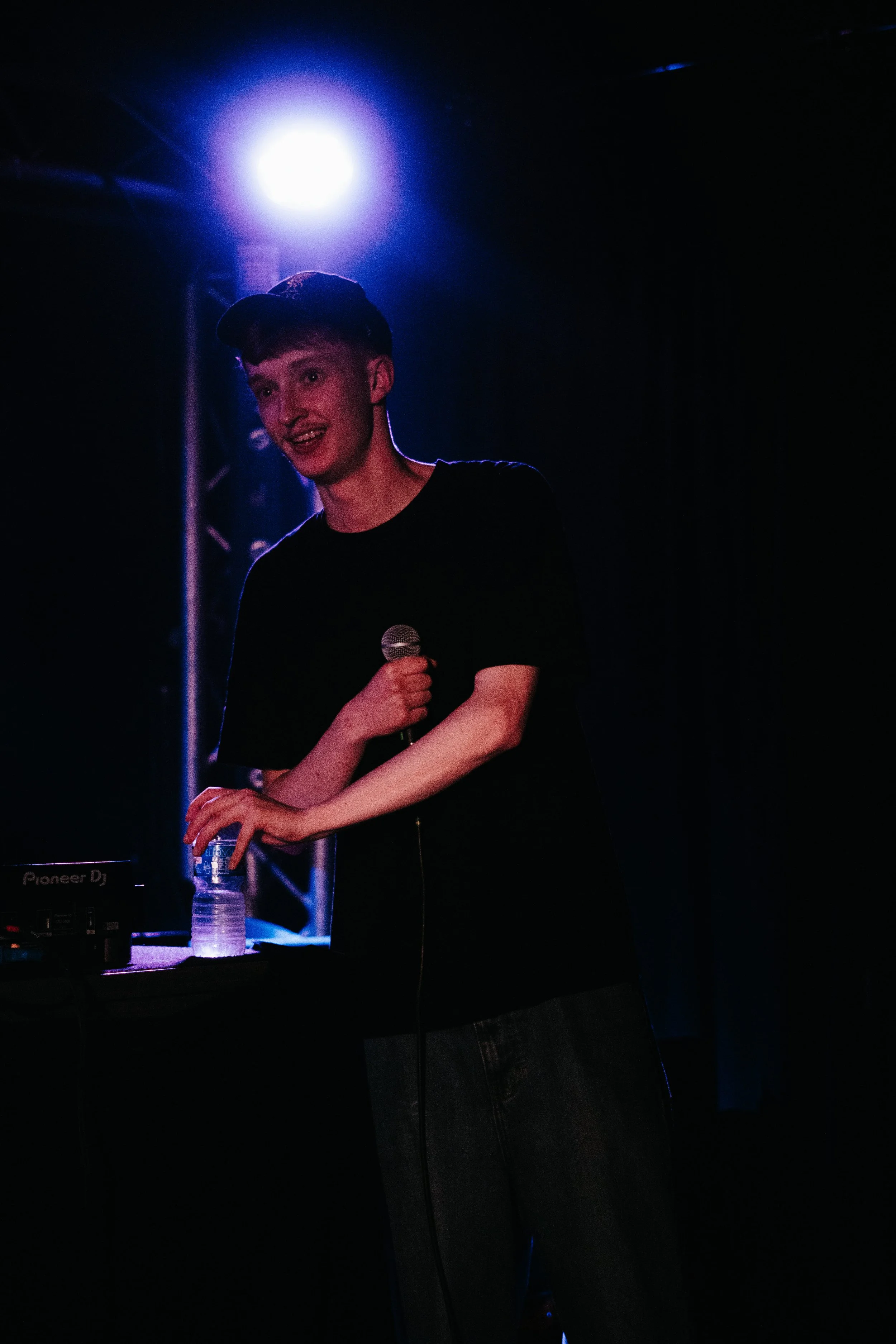 Jeune homme avec un chapeau noir, debout sur scène, tenant un microphone en main gauche, près d'une table avec un DJ et une bouteille d'eau, sous un éclairage violet