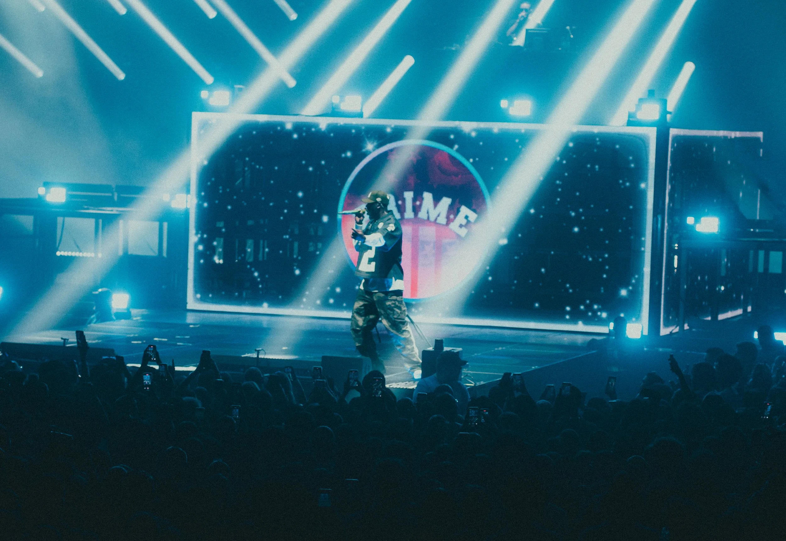 Un artiste en tenue décontractée avec un casque de réalité virtuelle, se produisant sur scène lors d'un concert ou d'un spectacle, avec un fond lumineux et un écran géant affichant un logo ou un nom, entouré par un public nombreux.