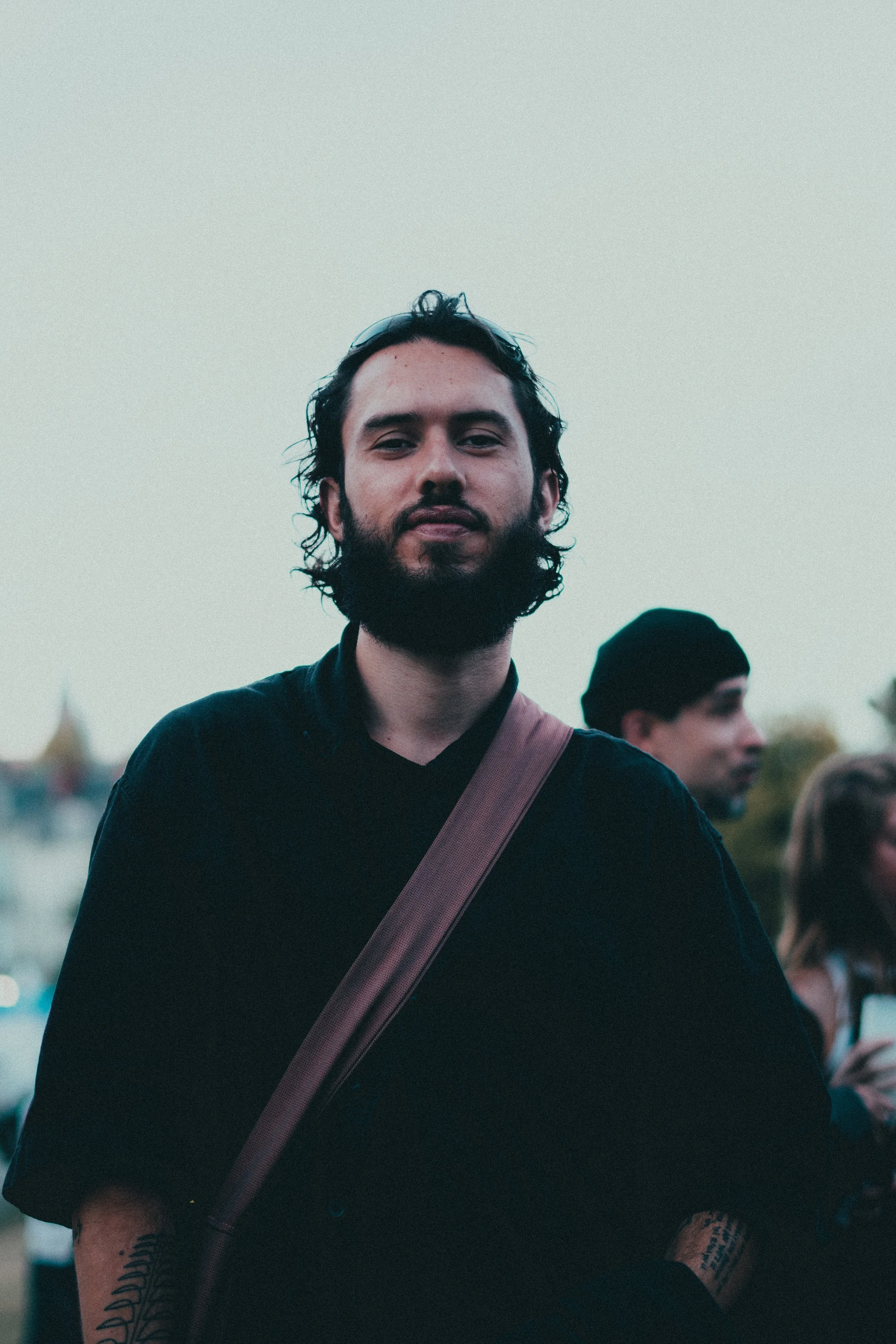 Un homme avec une barbe et des cheveux bouclés, portant un sac en bandoulière, se tient à l'extérieur avec un fond flou de plusieurs personnes.