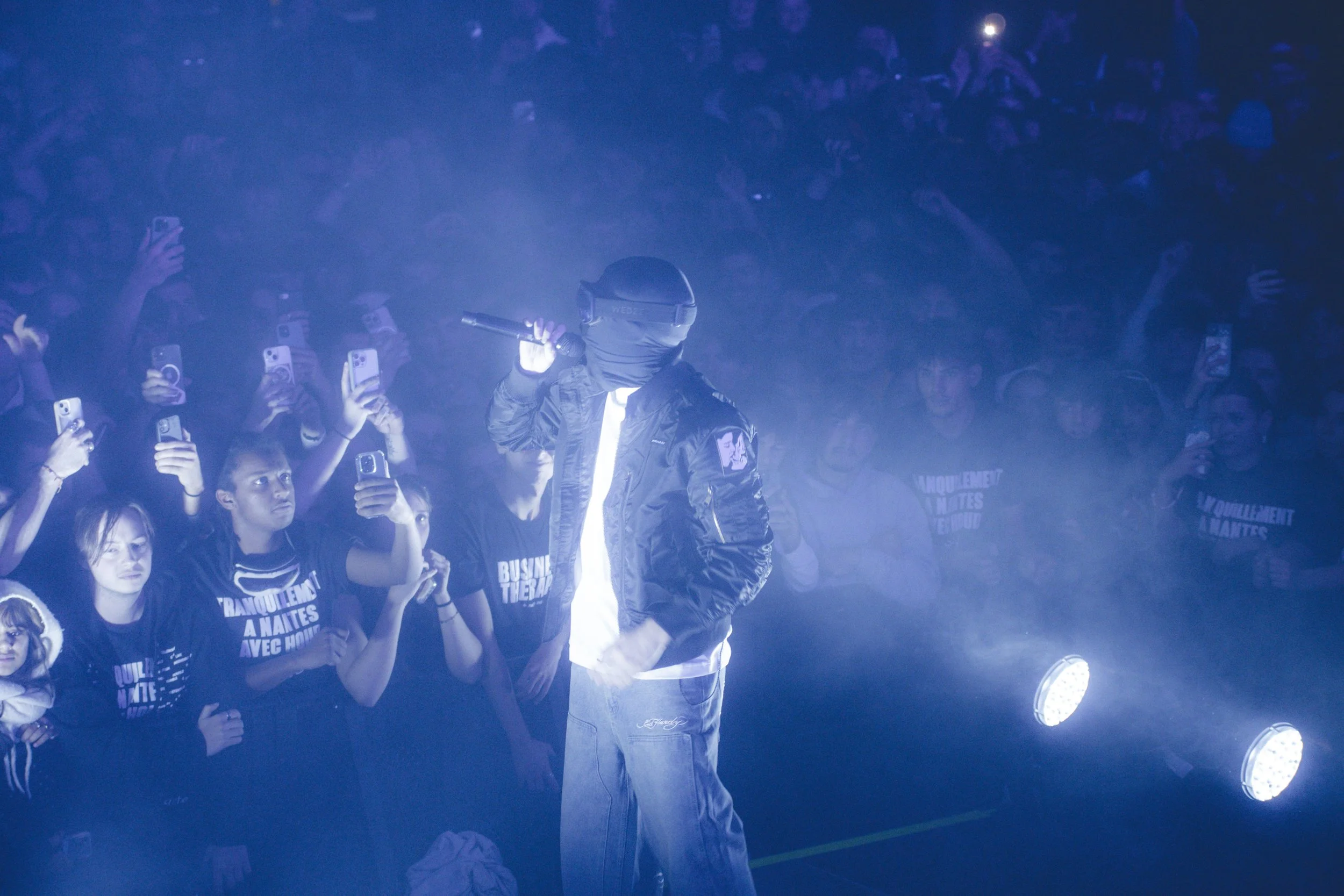 Une scène de concert ou d'événement avec un artiste portant un masque et des lunettes de réalité virtuelle, entouré d'un public prenant des photos ou vidéos avec leurs téléphones, dans un environnement sombre avec des lumières brillantes.