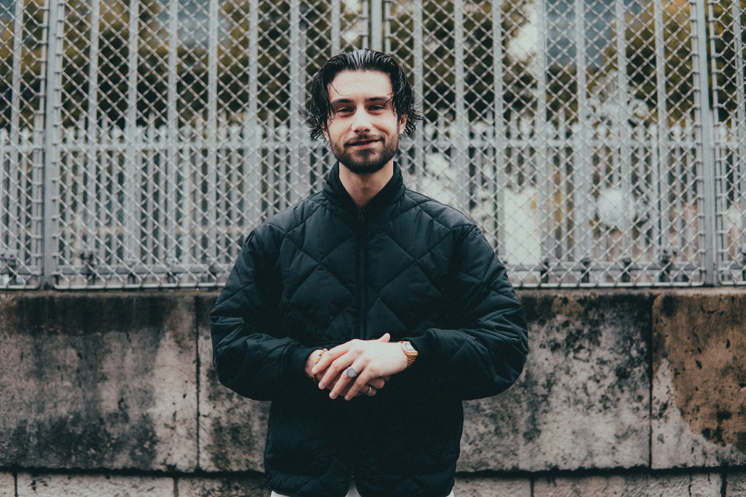 Jeune homme avec des cheveux noirs ondulés, portant une veste noire, posant devant une clôture en métal dans un cadre urbain.