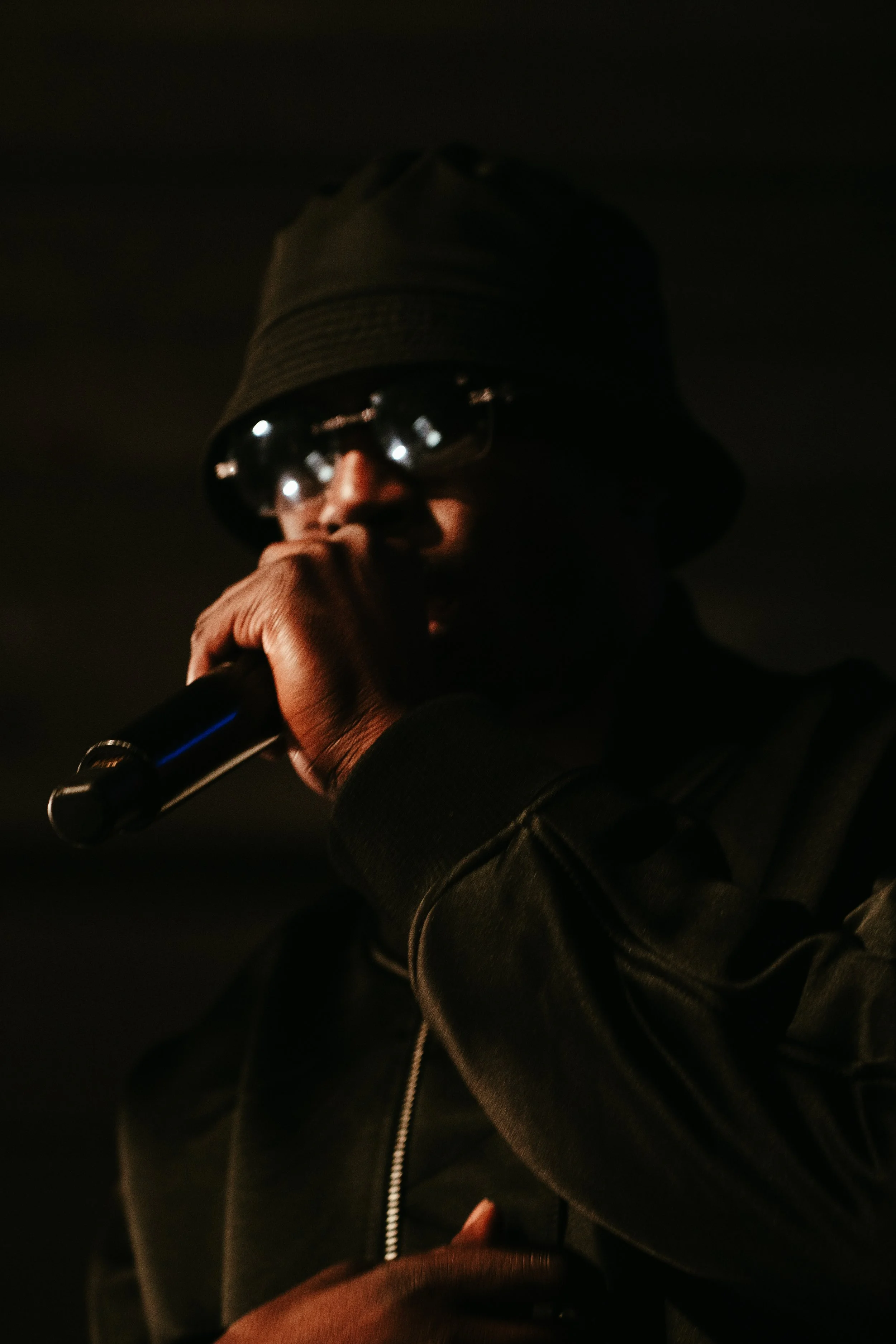 Un homme portant un chapeau, des lunettes de soleil et un blouson noir, tenant un microphone en train de chanter ou de parler dans un environnement sombre.