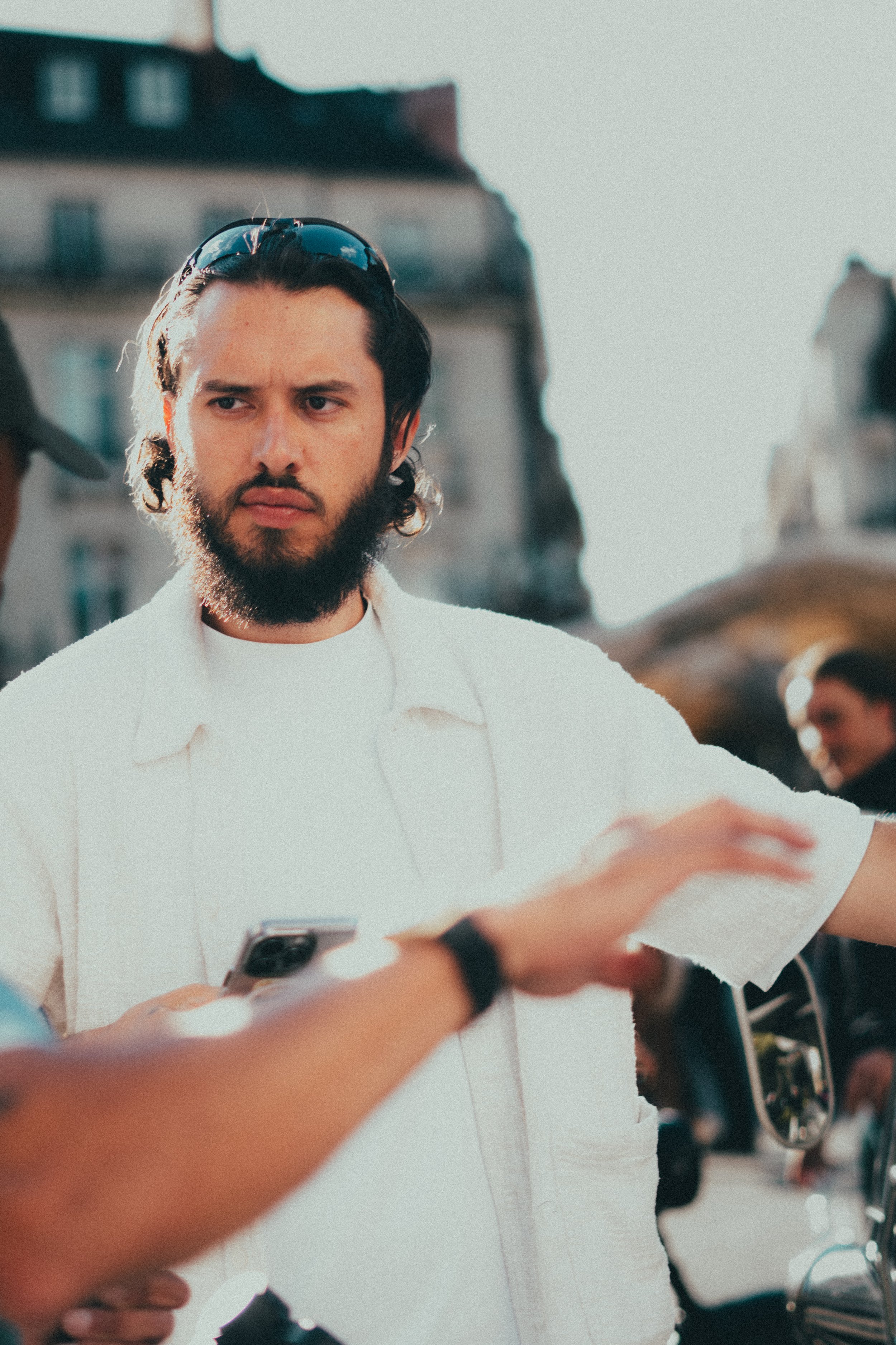 Un homme avec une barbe et des cheveux longs, portant une chemise blanche, tient un téléphone portable. Il semble écouter ou parler à quelqu'un lors d'une interaction en extérieur, dans une zone urbaine, avec d'autres personnes et bâtiments en arrièr