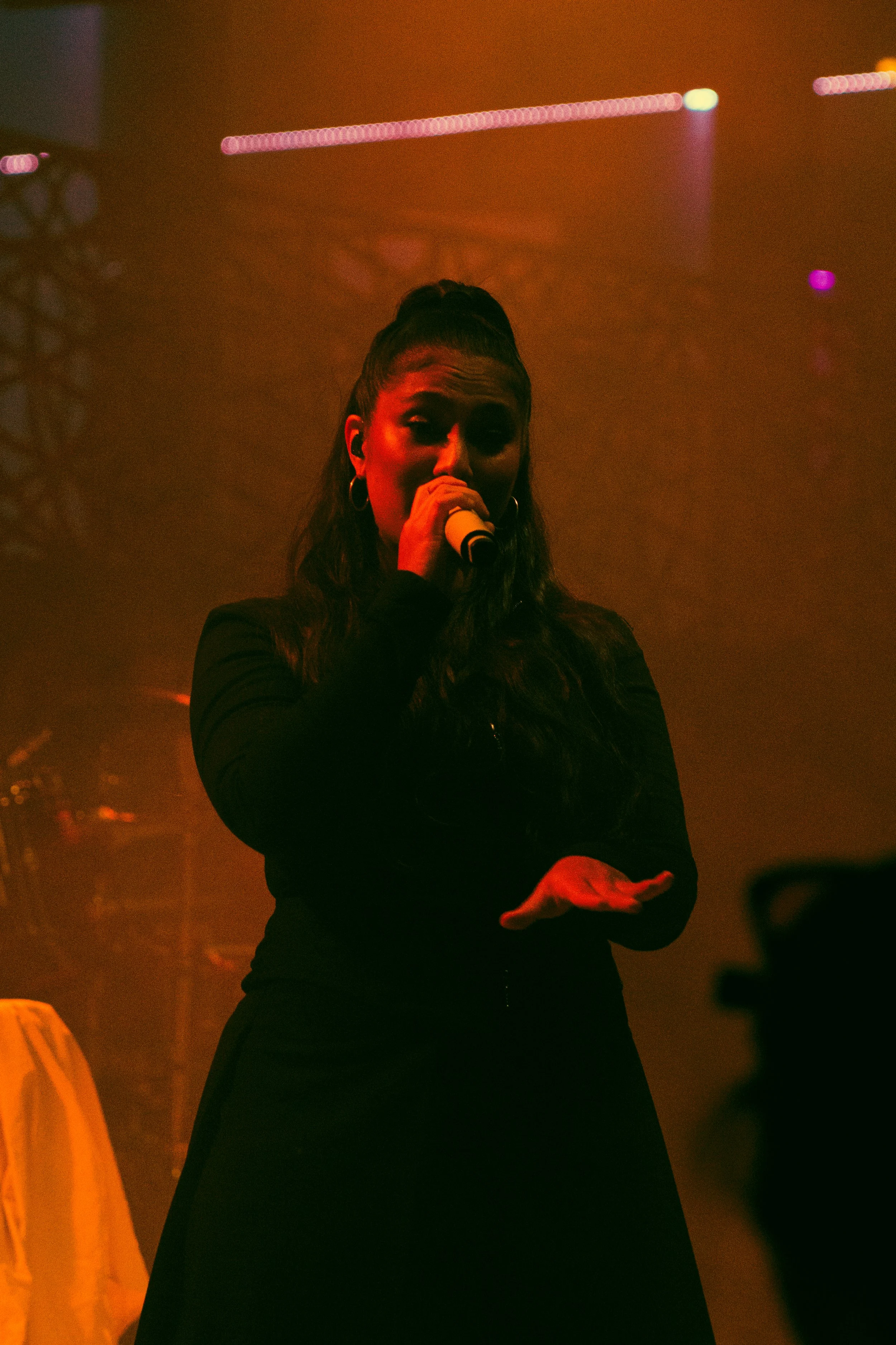 Une femme chante avec un micro dans une scène sombre éclairée par des lumières colorées, avec une atmosphère de concert.