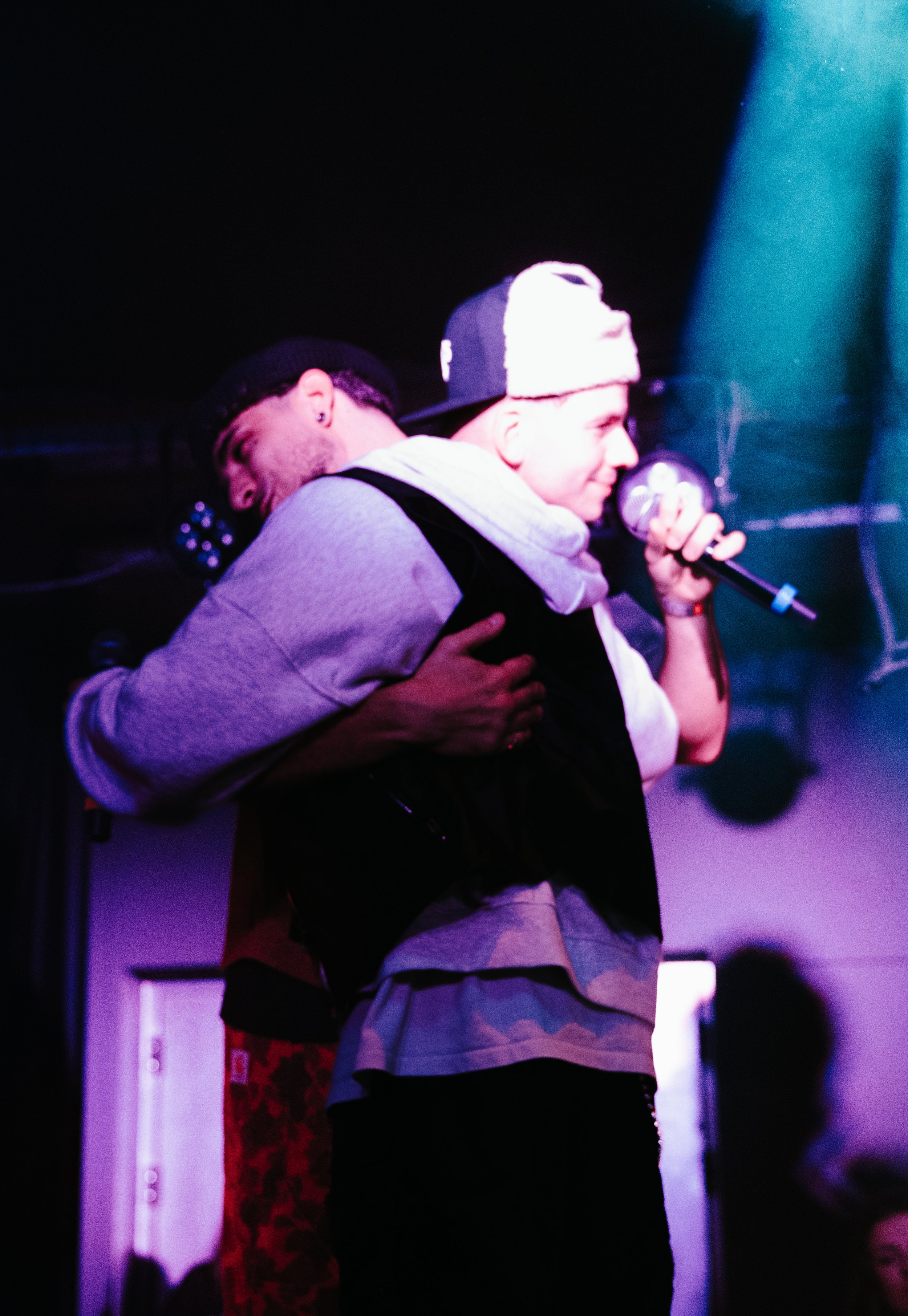 Deux jeunes hommes s'étreignent sur scène, l'un portant un bonnet noir, et l'autre un bonnet blanc, tenant un micro, dans une ambiance de spectacle ou de concert.