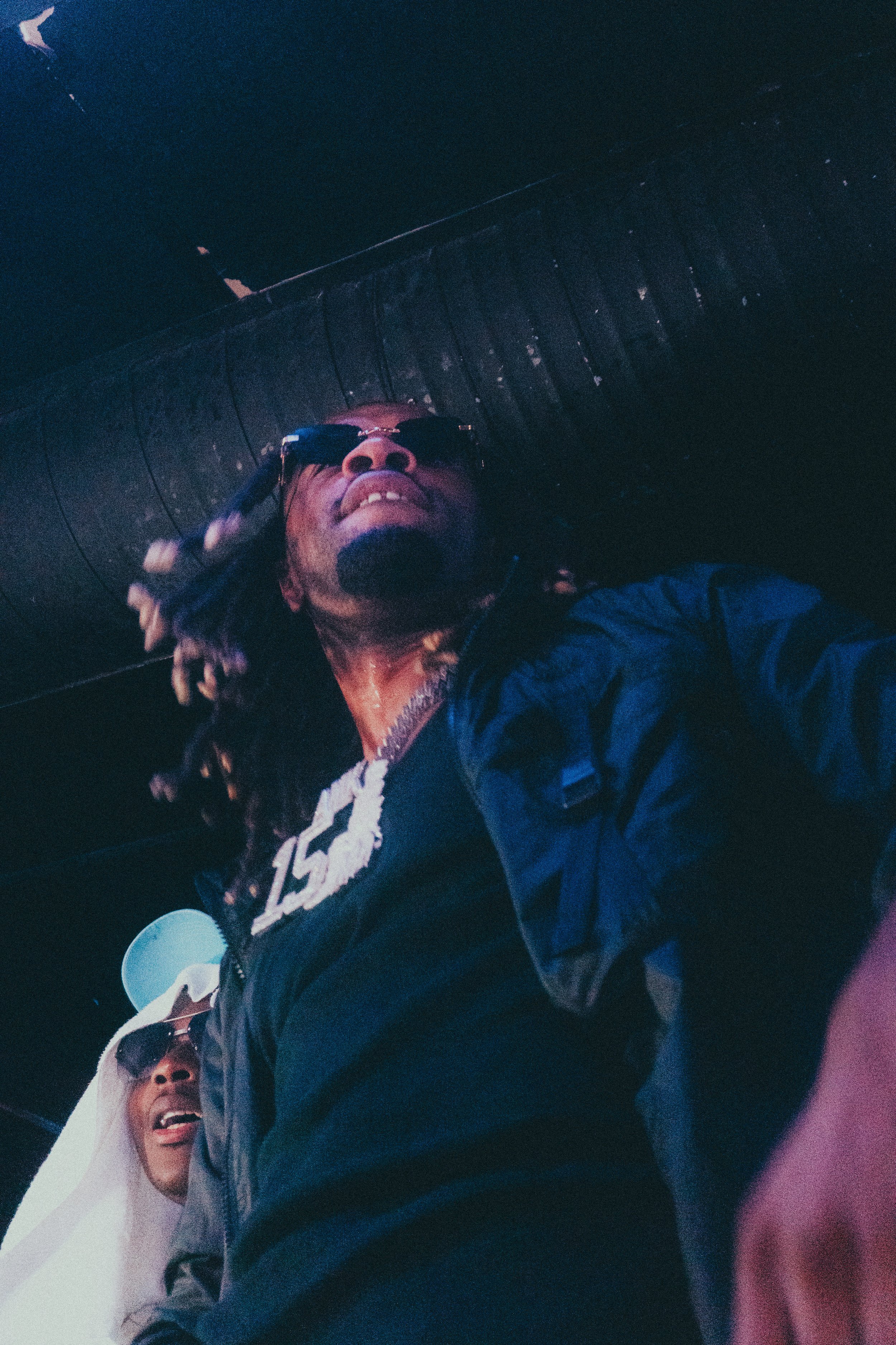 Deux hommes portant des lunettes de soleil et des vêtements décontractés, en train de poser pour une photo dans un décor sombre.