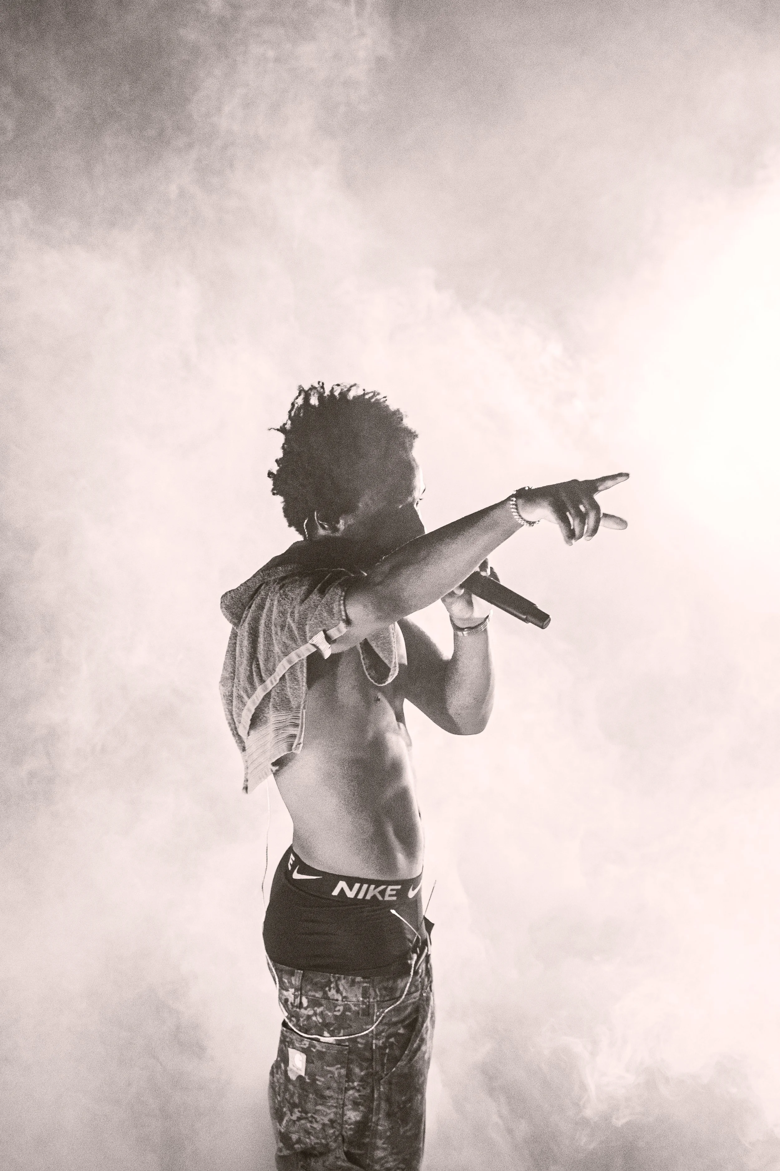 Un homme nu, portant un pantalon de survêtement Nike, tenant un micro et pointant avec son doigt vers la droite, avec un vêtement sur son épaule.