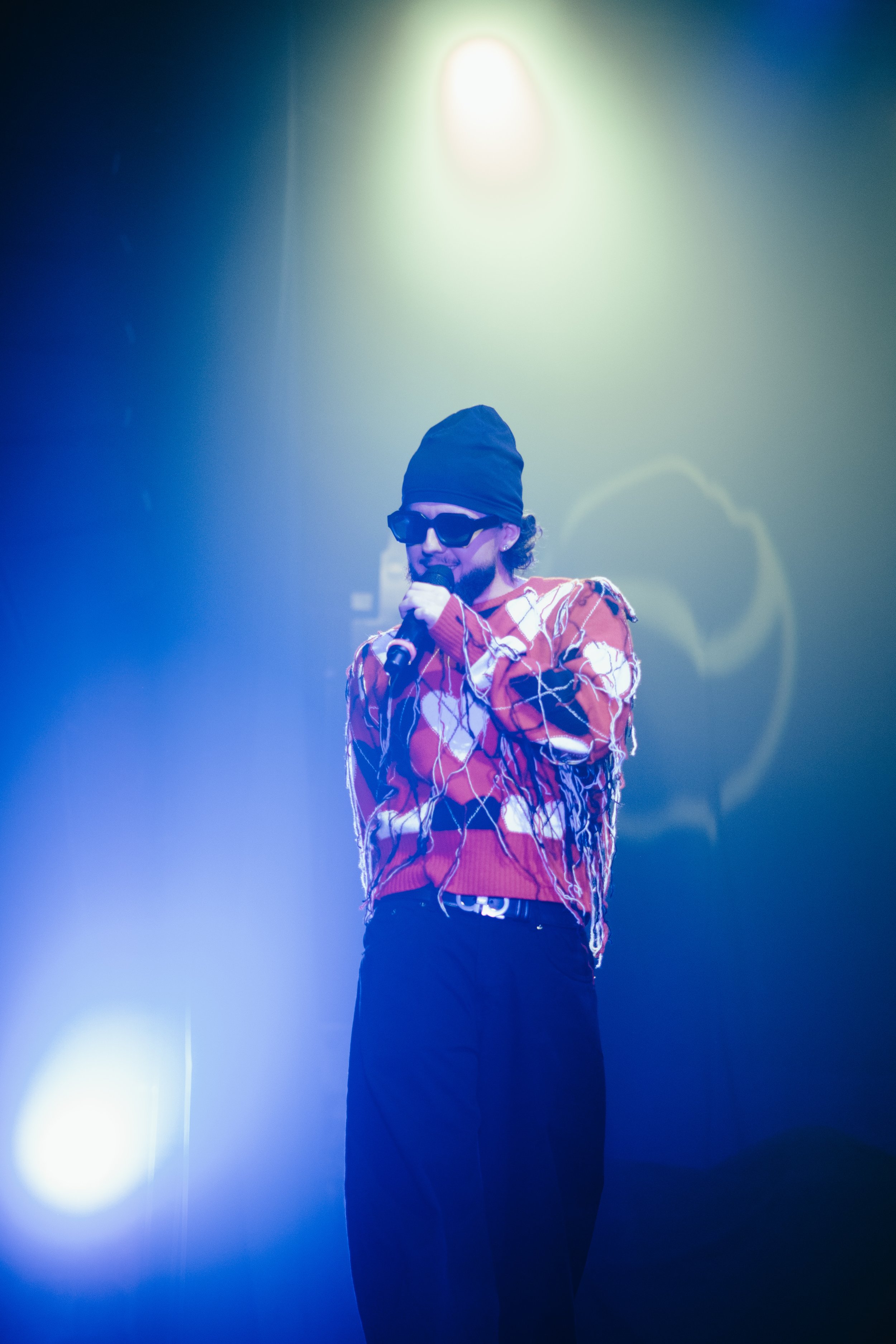 Un homme portant un bonnet noir, des lunettes de soleil et un pull rouge à motifs blancs, chantant dans un micro, avec un éclairage de scène coloré.