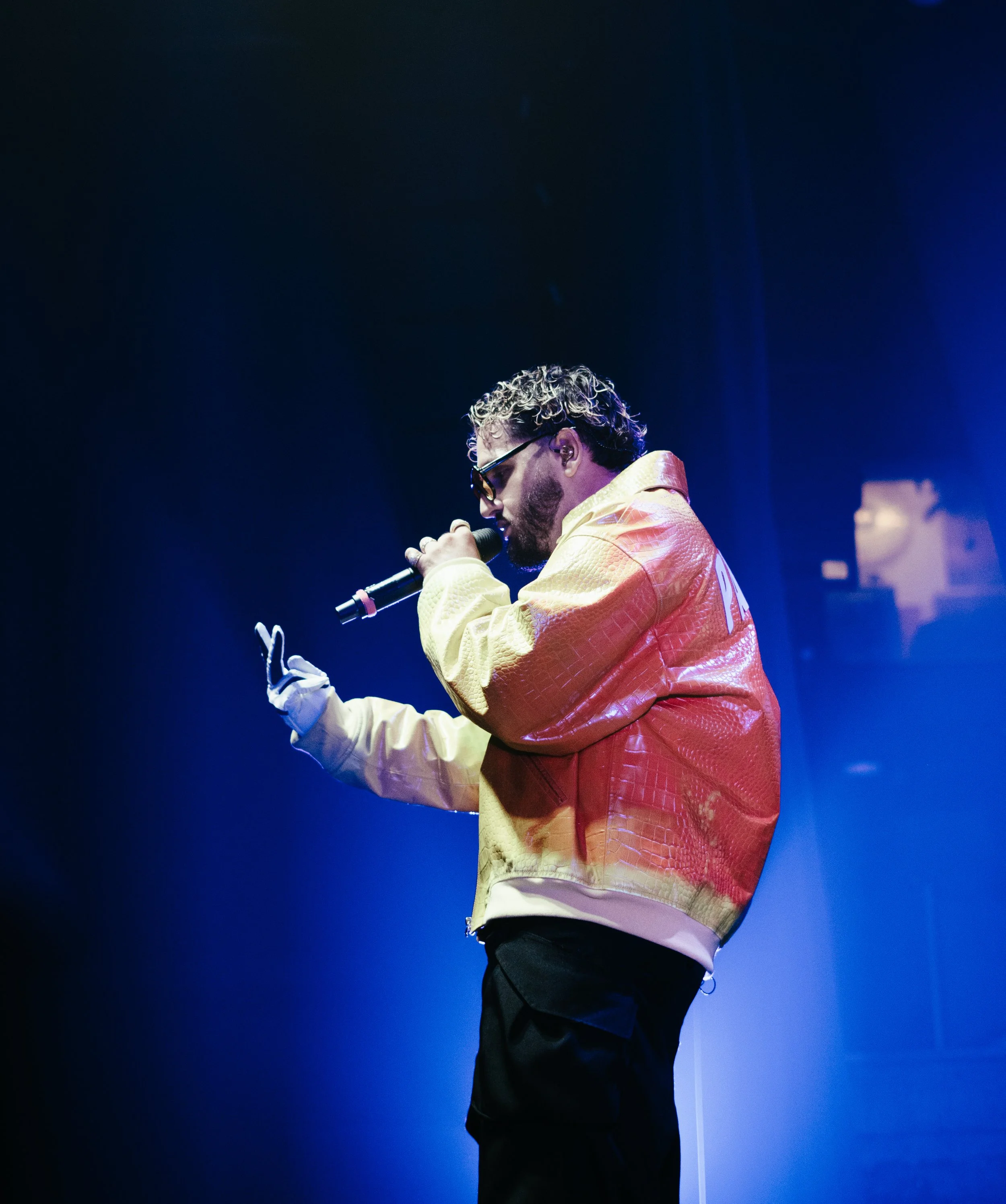 Un homme portant des lunettes de soleil, une veste jaune et rouge, tient un microphone et semble chanter ou rapper sur scène, avec un éclairage bleu en arrière-plan.