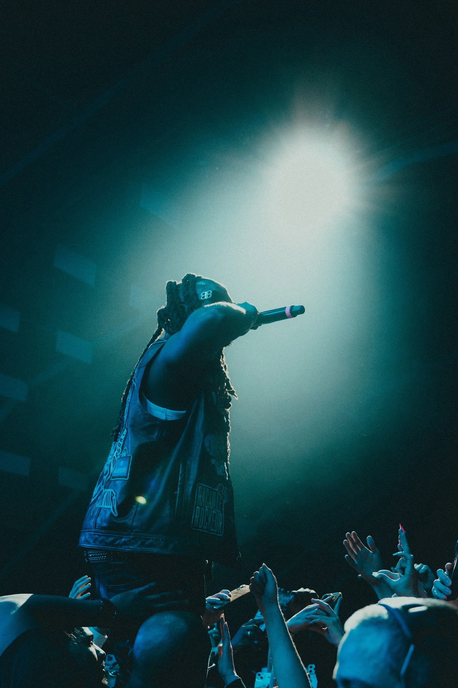 Un artiste de rap sur scène, tenant un micro, éclairé par un projecteur, avec des mains levées dans le public.