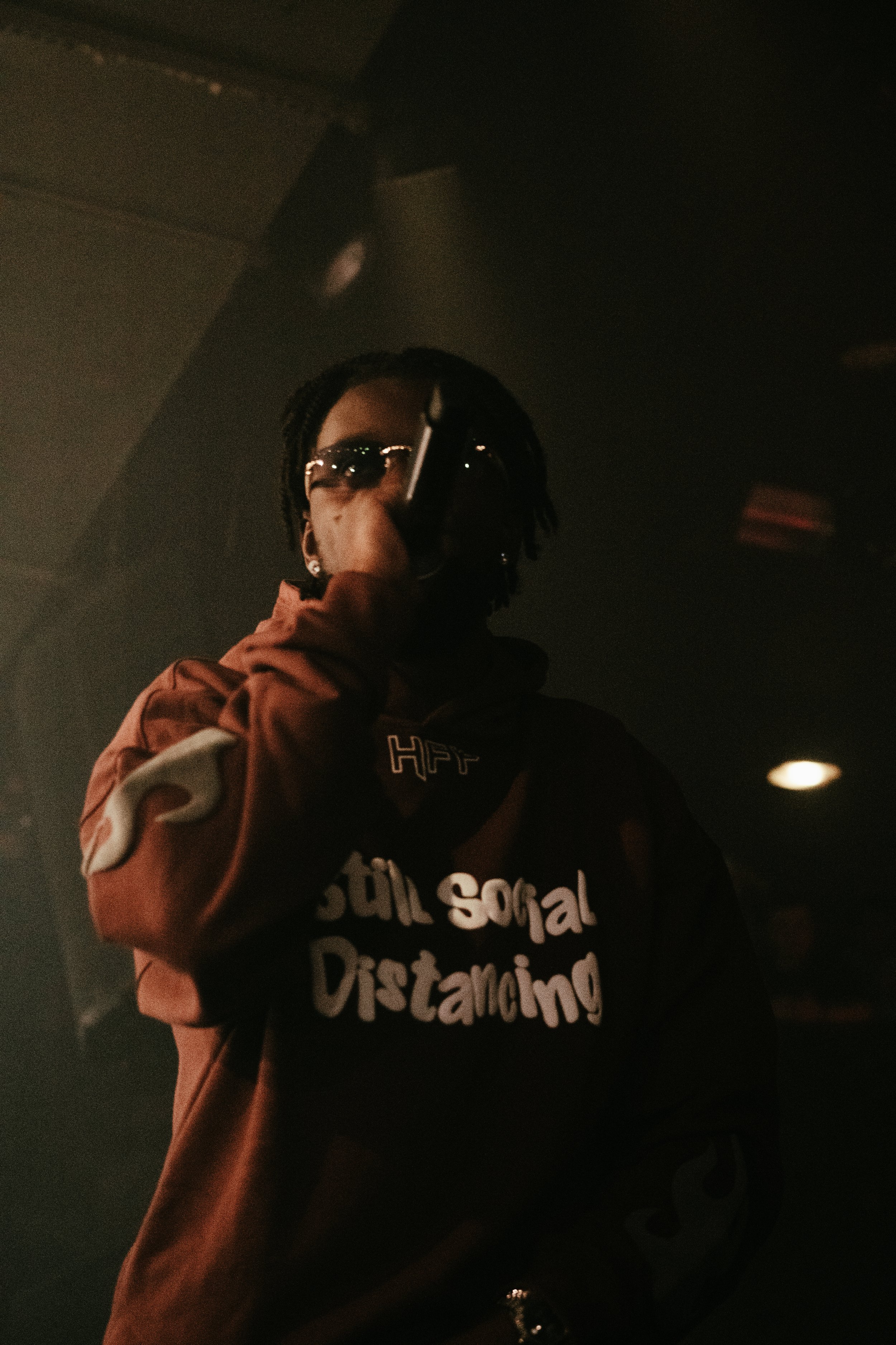 Une personne portant une veste marron avec le texte 'Still Social Distancing' en blanc, portant des lunettes de soleil et un masque, dans un endroit sombre.