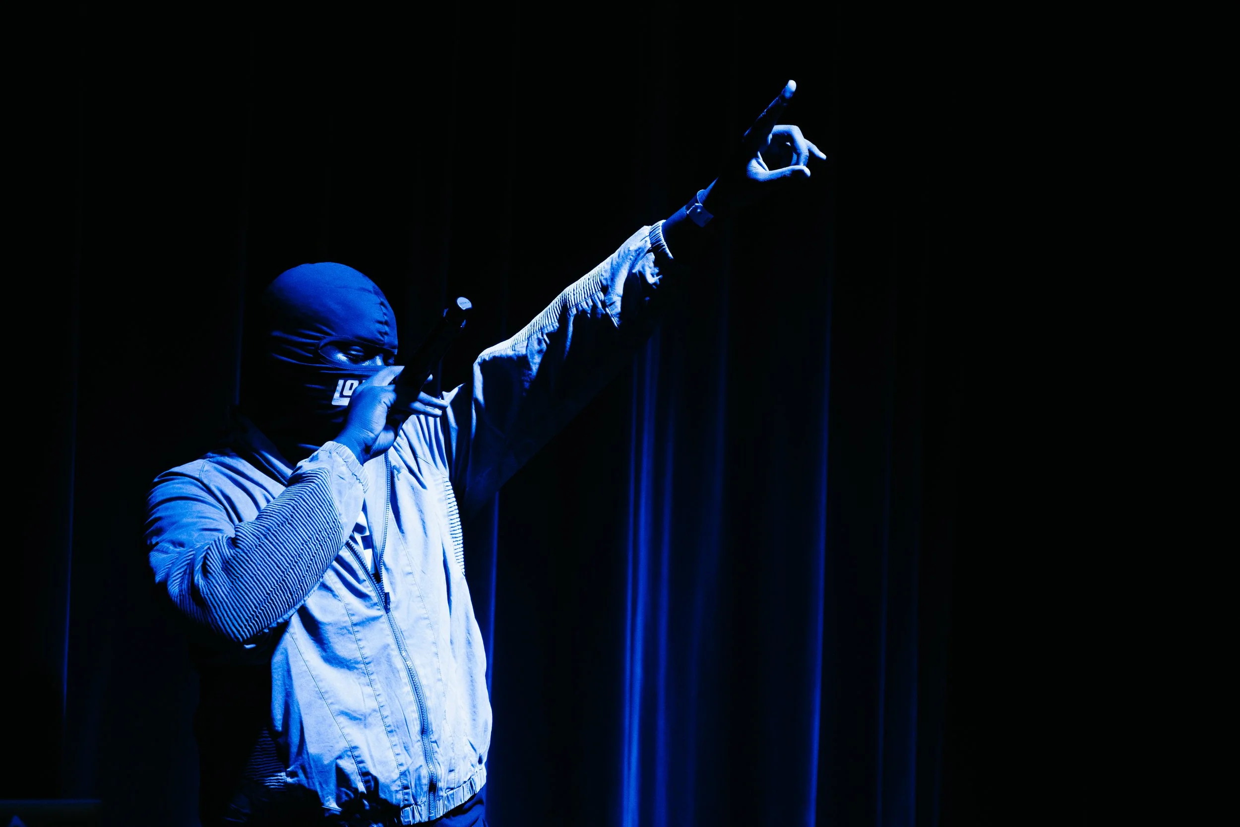 Une personne portant un masque noir, tenant un microphone et levant une main, éclairée par une lumière bleue dans une scène sombre.