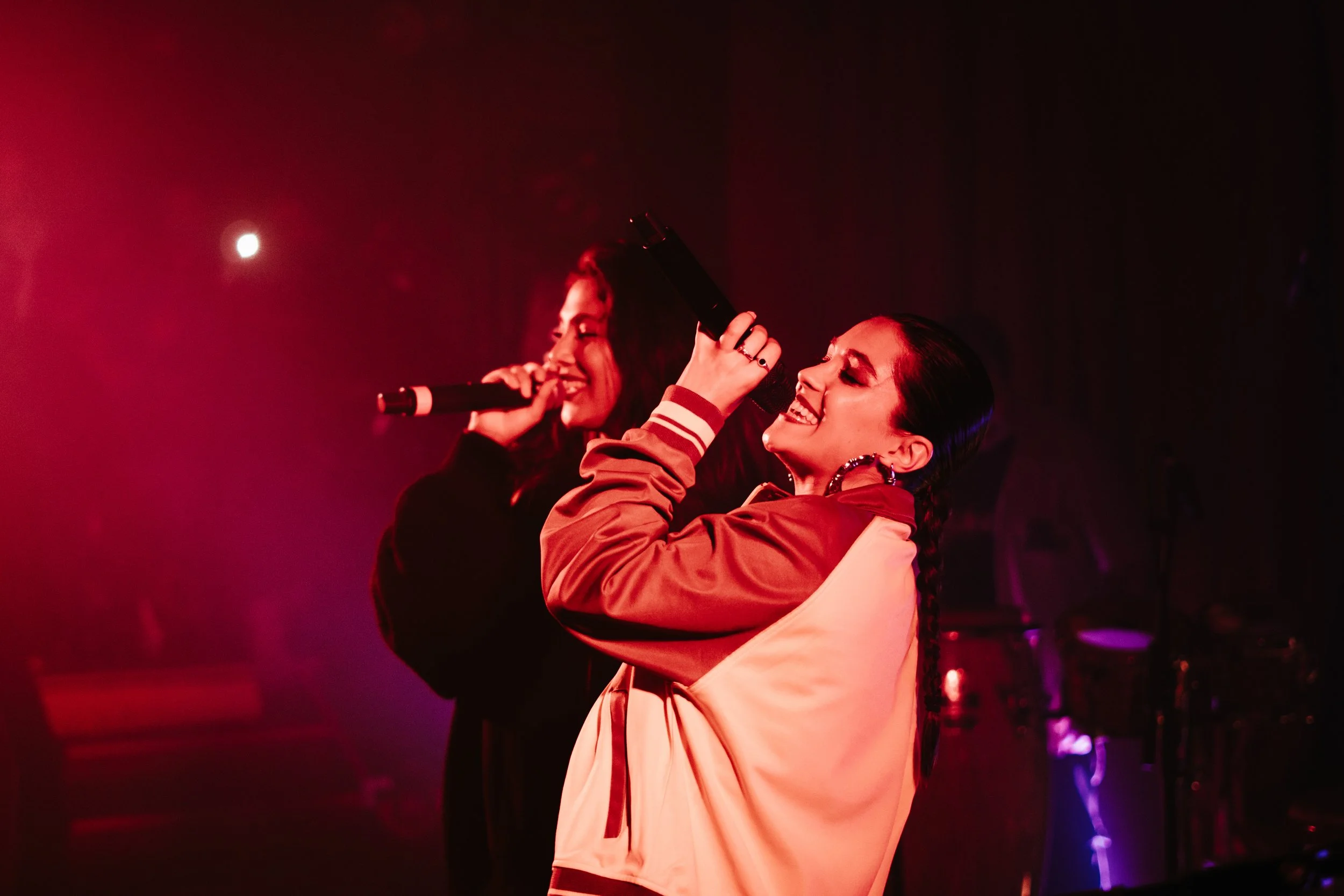 Deux femmes chantent sur scène sous un éclairage rouge, l'une en tenue de sport et l'autre en tenue noire, avec microphones à la main.