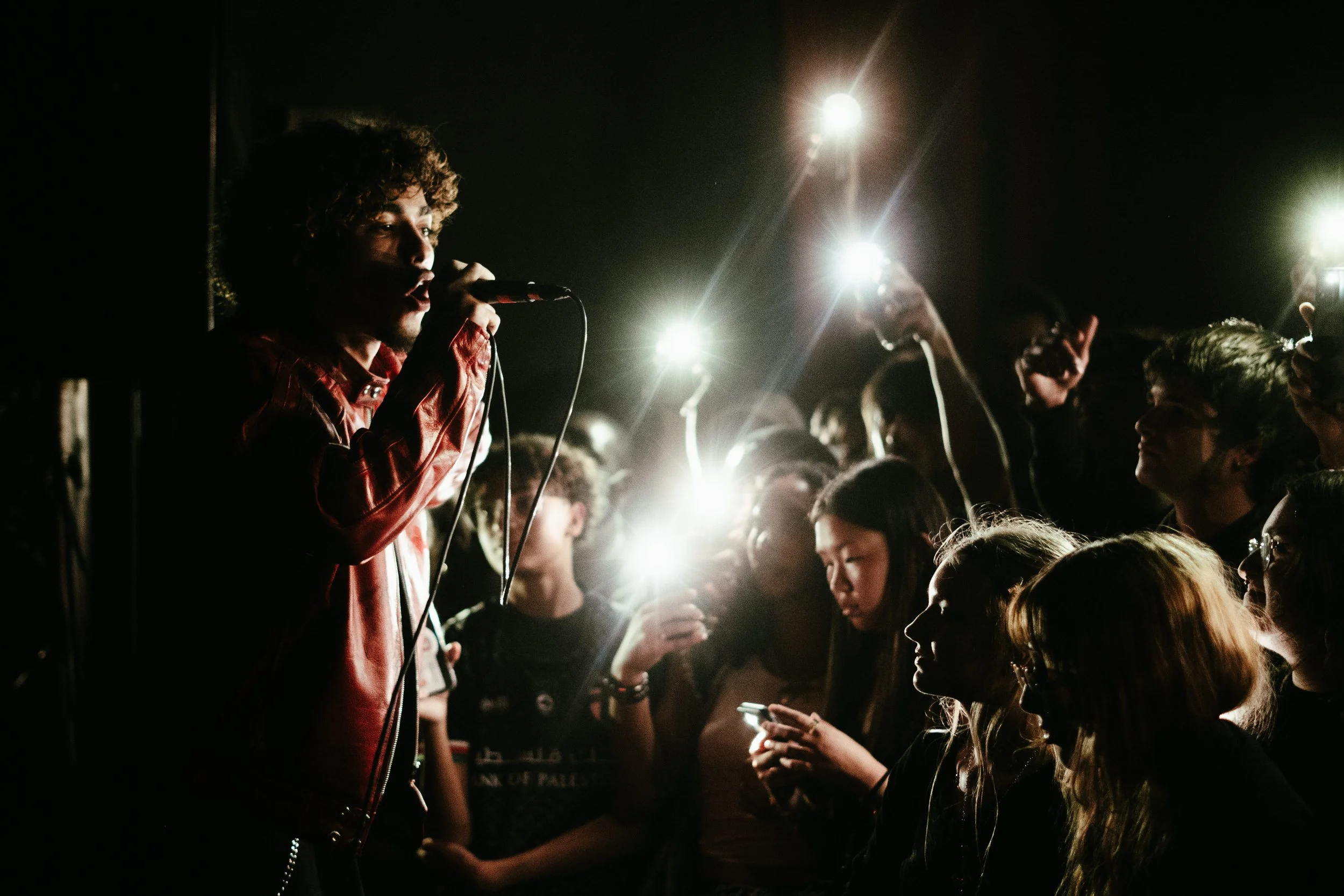 Un jeune homme chante avec un microphone lors d'un concert, entouré d’un public avec des lumières brillantes derrière eux.
