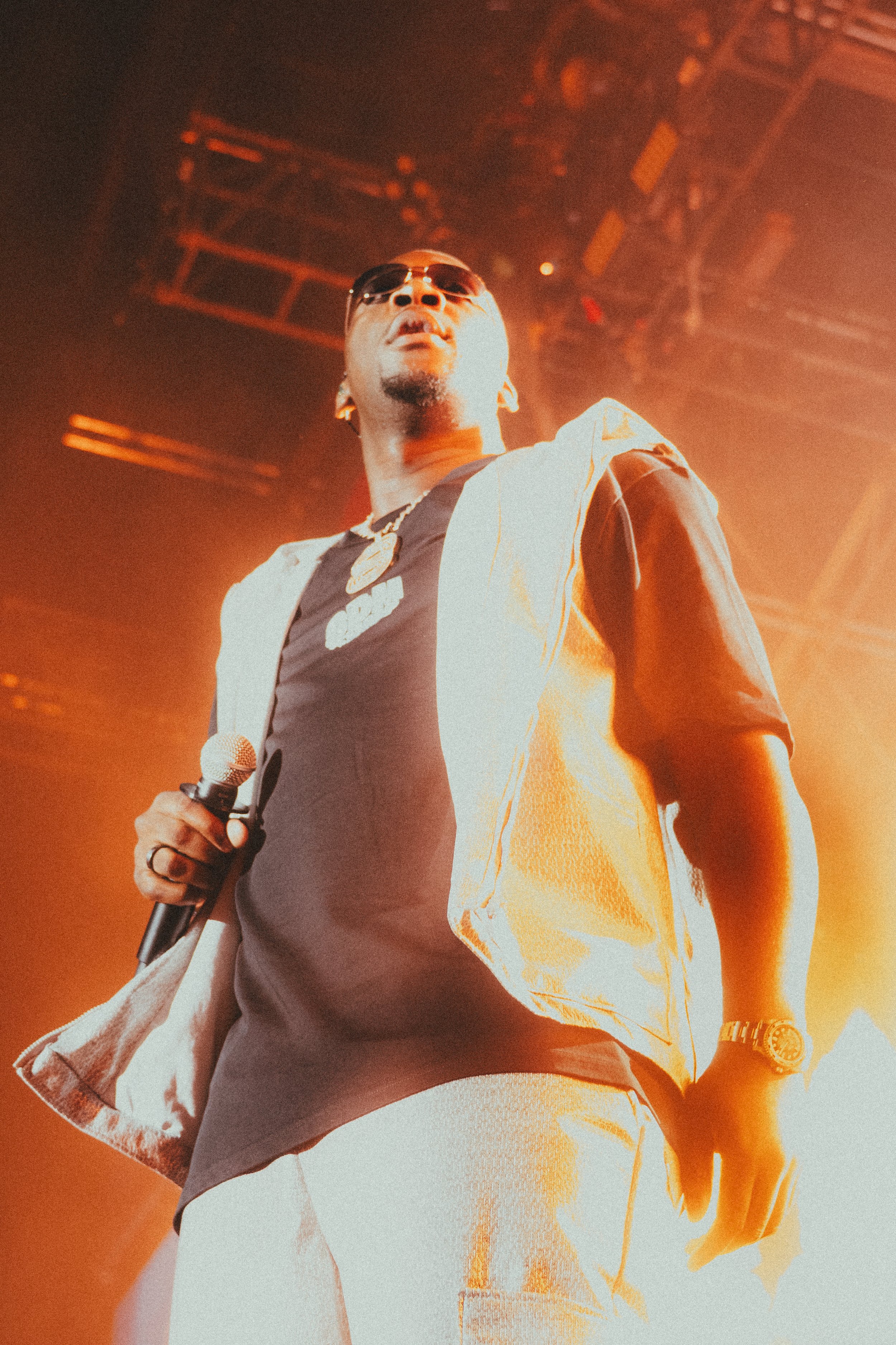Un homme portant des lunettes de soleil, une chaîne en or et une montre, tenant un micro, sur scène avec un éclairage chaud et ambiance de concert.