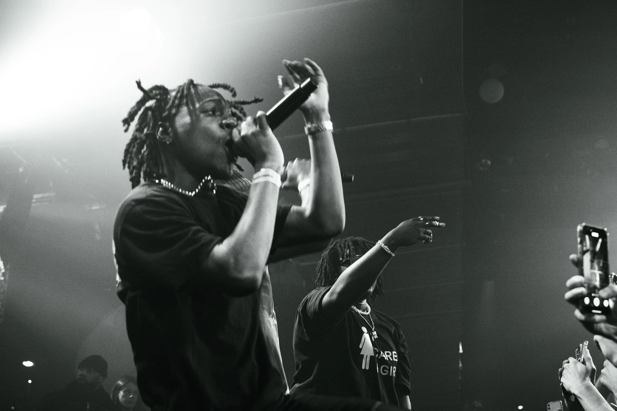 Une femme chante avec un microphone lors d'un concert, entourée d'un public. En arrière-plan, une autre personne lève la main.