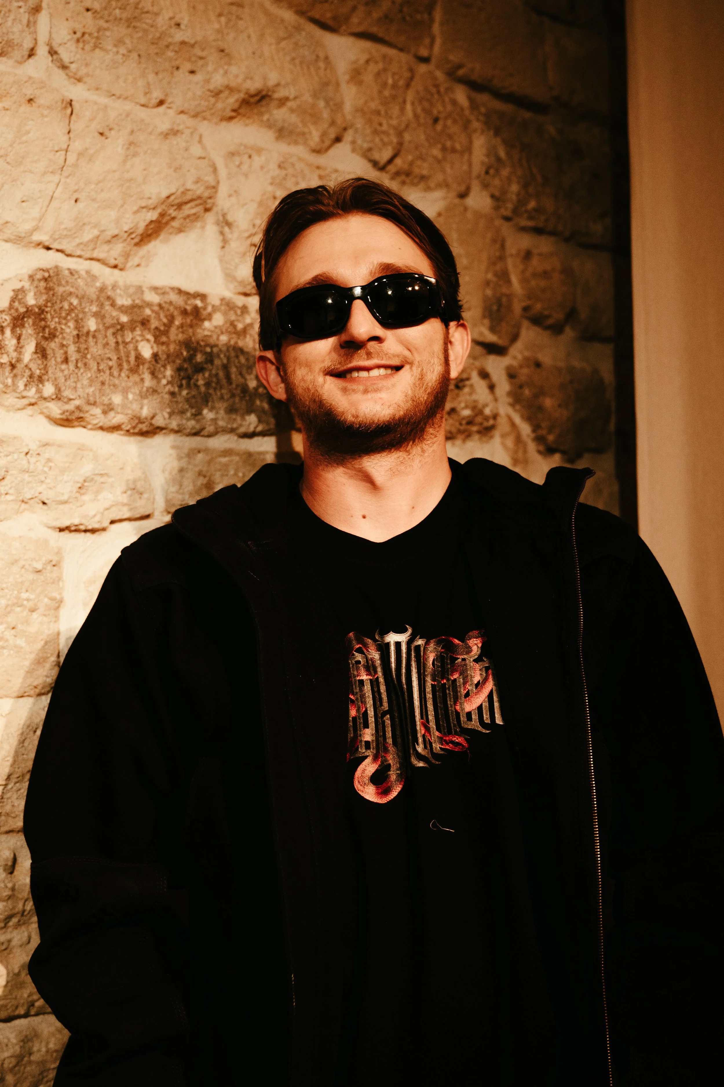 Jeune homme souriant portant des lunettes de soleil noires, en t-shirt noir avec un design en gothique, et une veste noire, posant devant un mur en pierre.