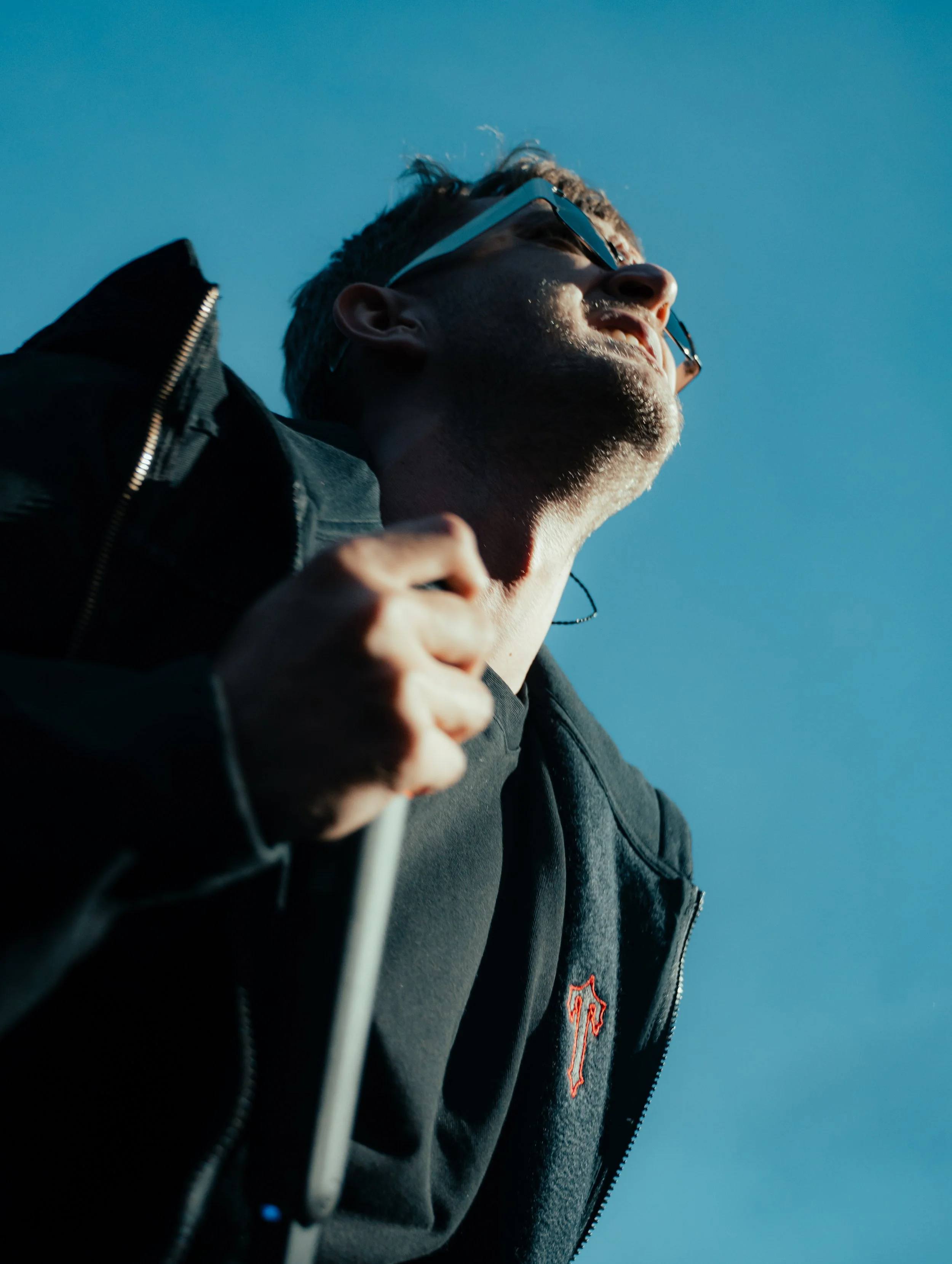 Jeune homme portant des lunettes de soleil et une veste noire, en train de courir ou marcher sous un ciel bleu clair.