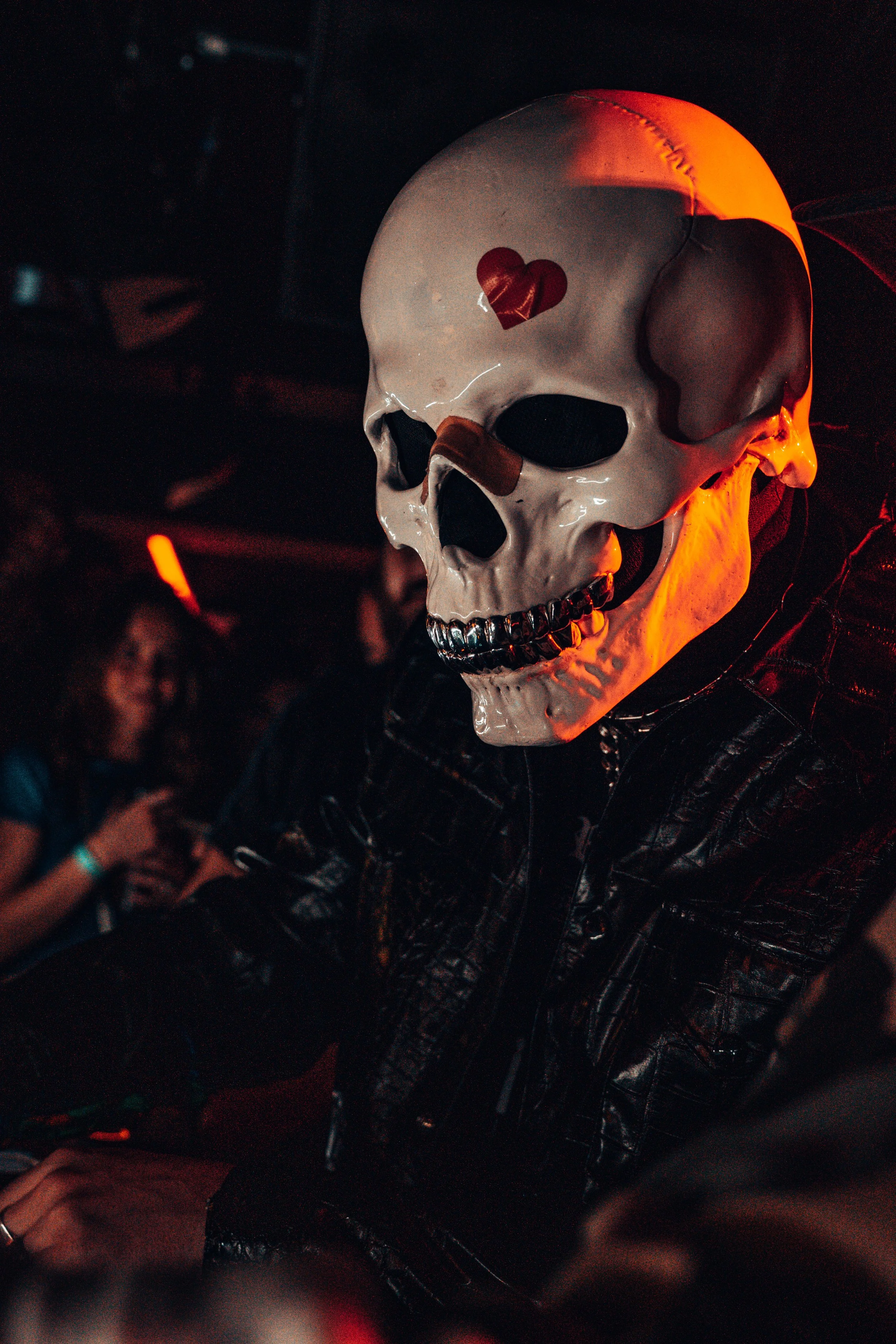 Une personne portant un masque de squelette avec un cœur découpé sur le front, vêtue d'une veste en cuir noire, dans un environnement sombre avec des lumières rouges.