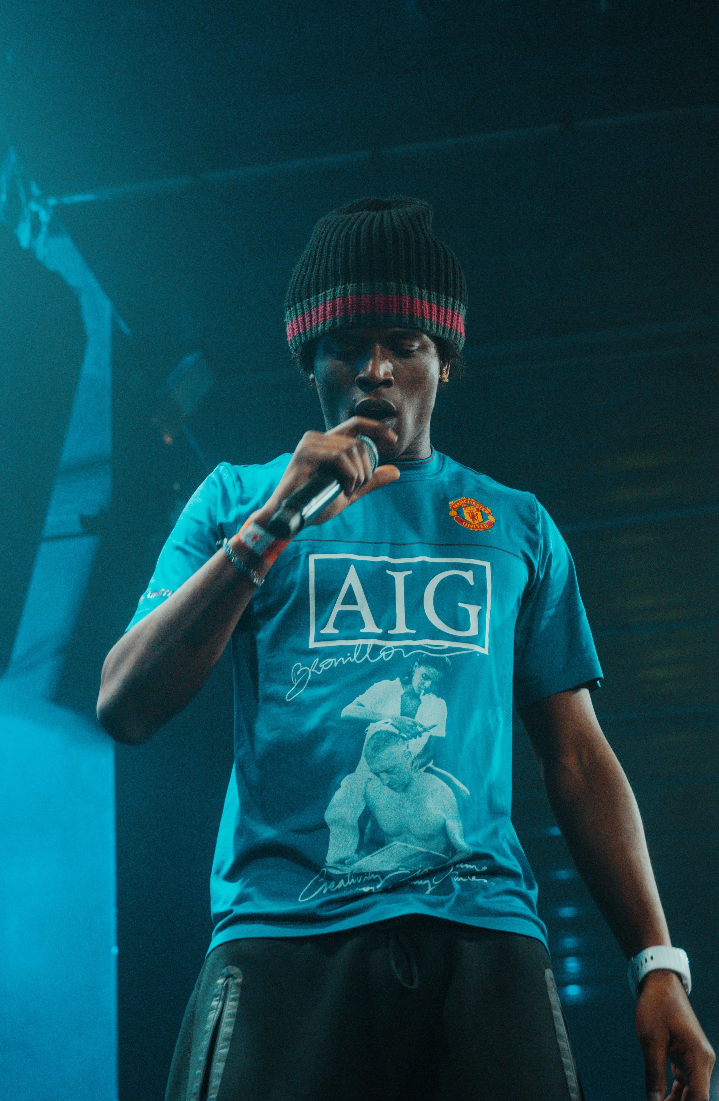 Une personne portant un sweat à capuche bleu avec un logo d'une équipe de football et une illustration, parlant dans un microphone, portant un bonnet et un bracelet blanc, dans un environnement sombre et éclairé par des lumières colorées.