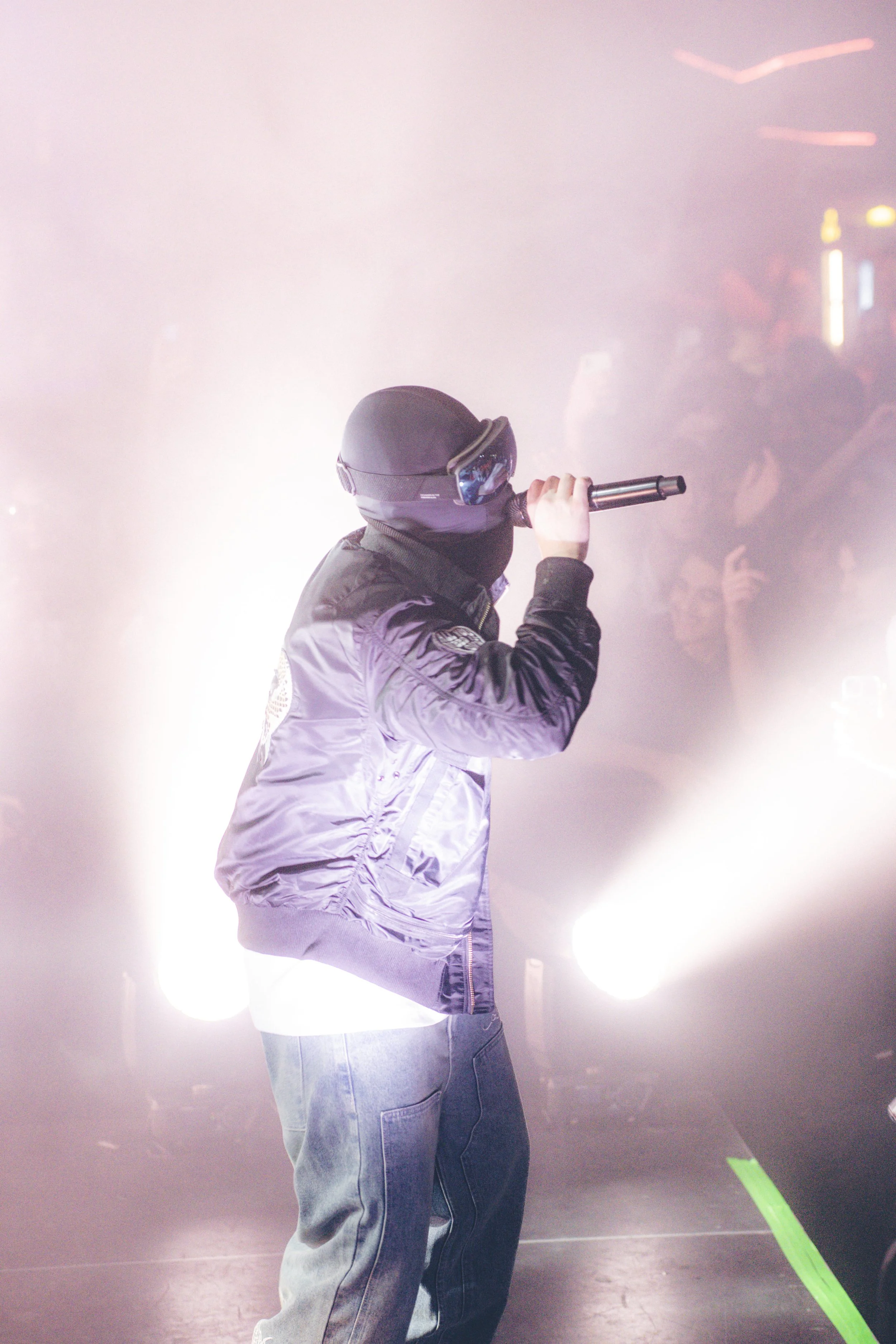 Un artiste de rap portant un casque noir et une veste bomber noire, tenant un microphone sur scène avec des lumières aveuglantes et un public en arrière-plan.