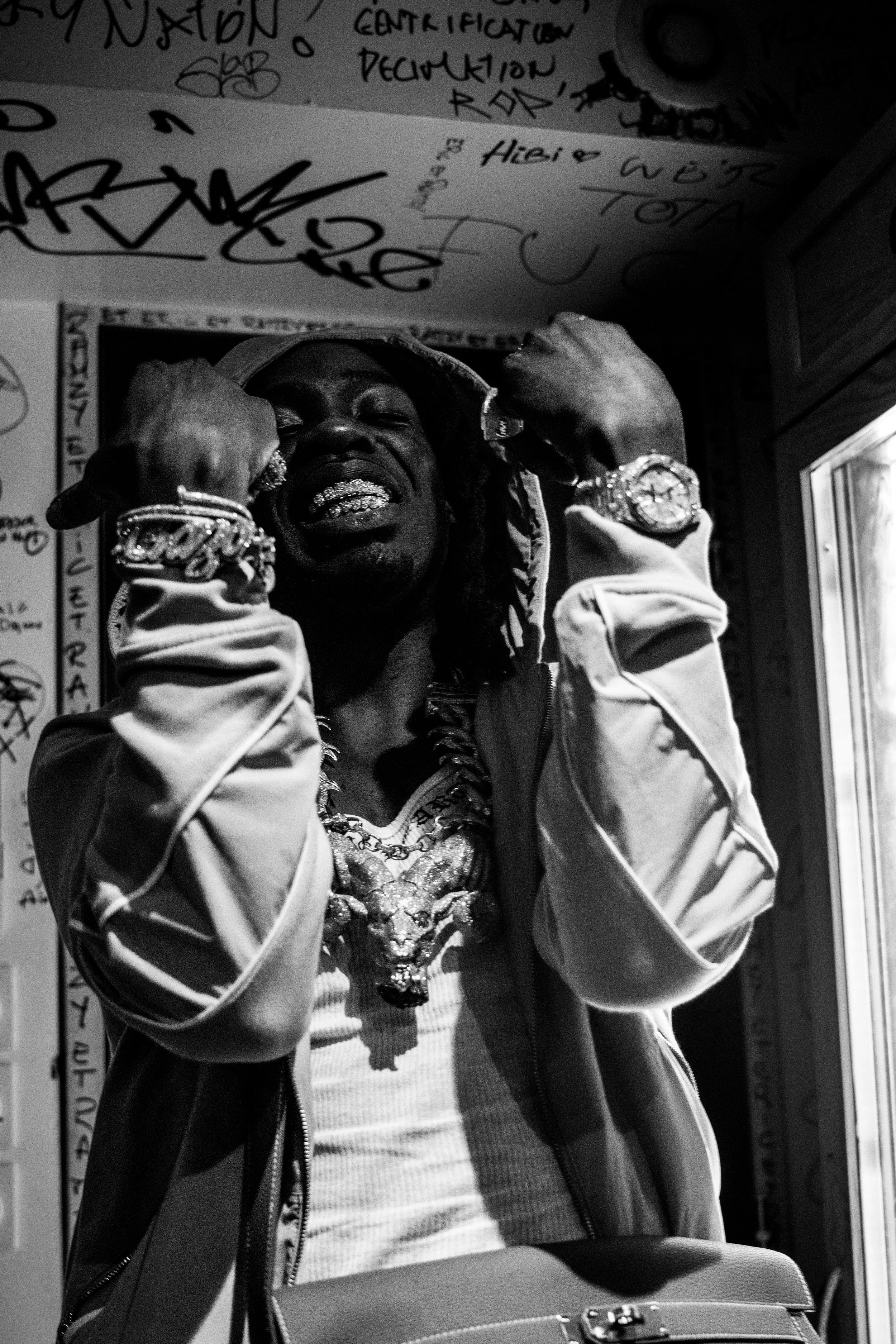 Un homme portant des bijoux voyants et une veste, le visage exprimant une émotion intense, se tient dans une pièce avec des graffitis sur les murs.