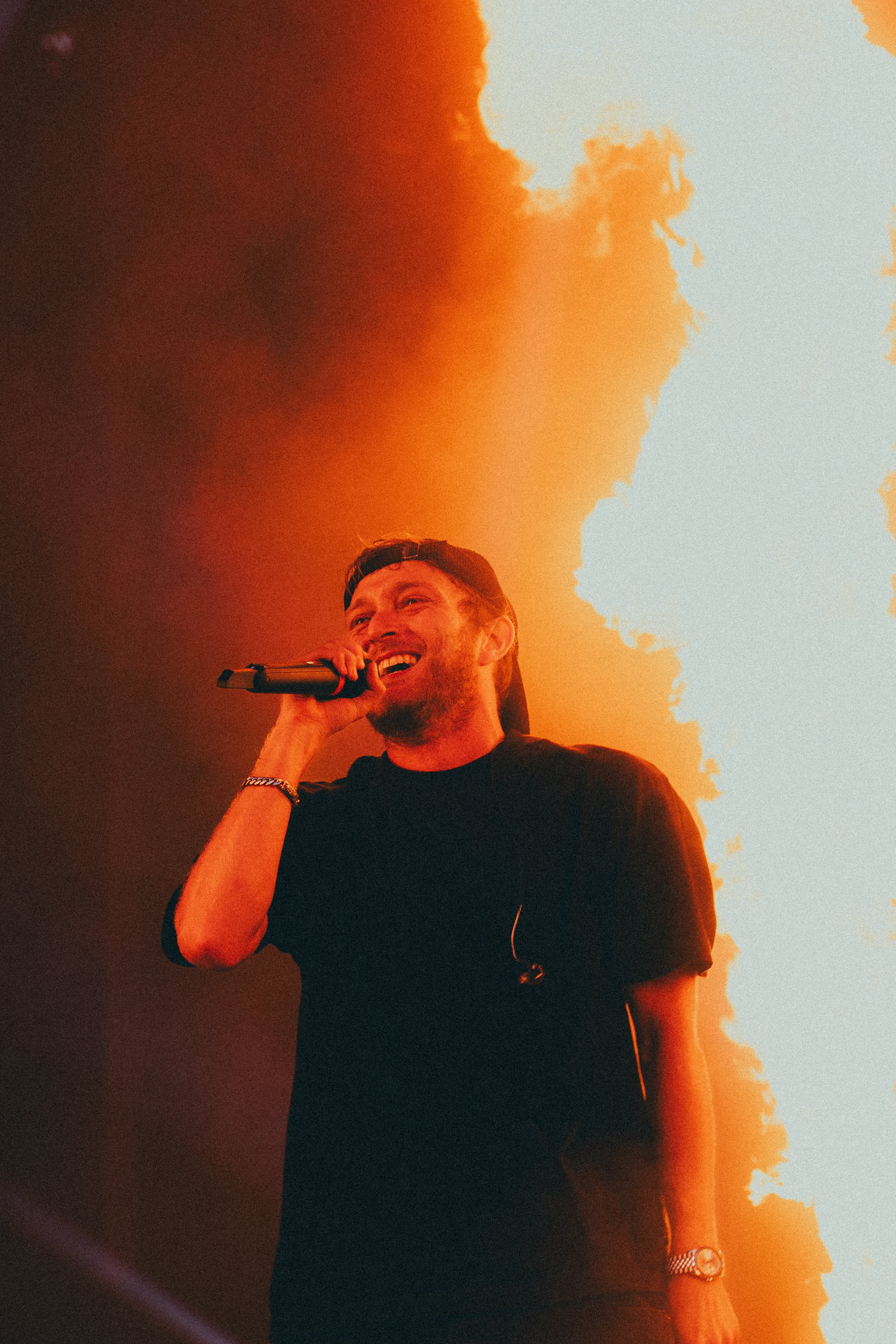 Un homme souriant tenant un microphone lors d'une performance sur scène avec un arrière-plan lumineux orange et blanc.