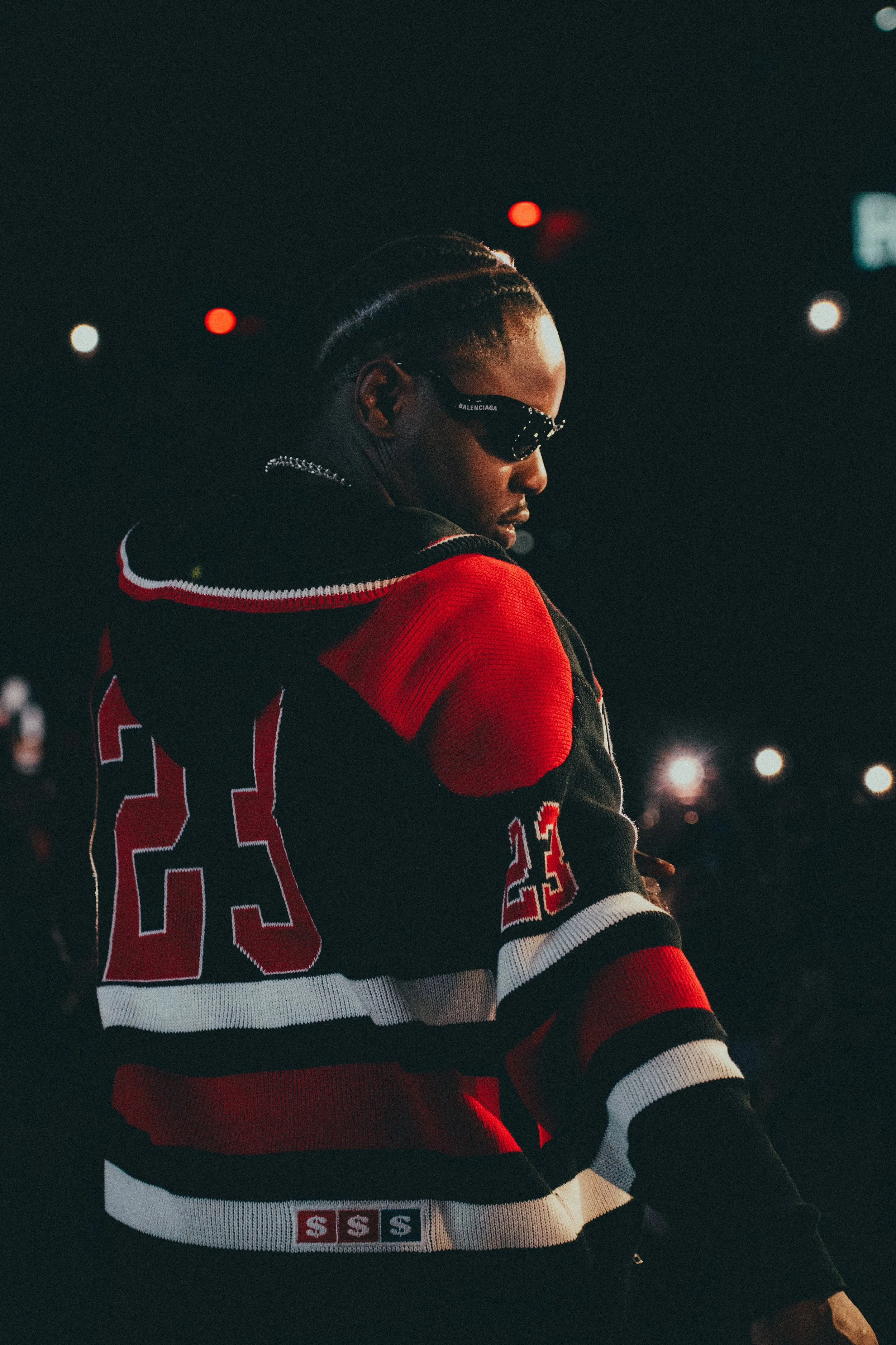 Une femme portant des lunettes de soleil noires, une veste à rayures rouges, noires et blanches avec le numéro 23 et un logo. Il fait sombre, avec des lumières en arrière-plan.