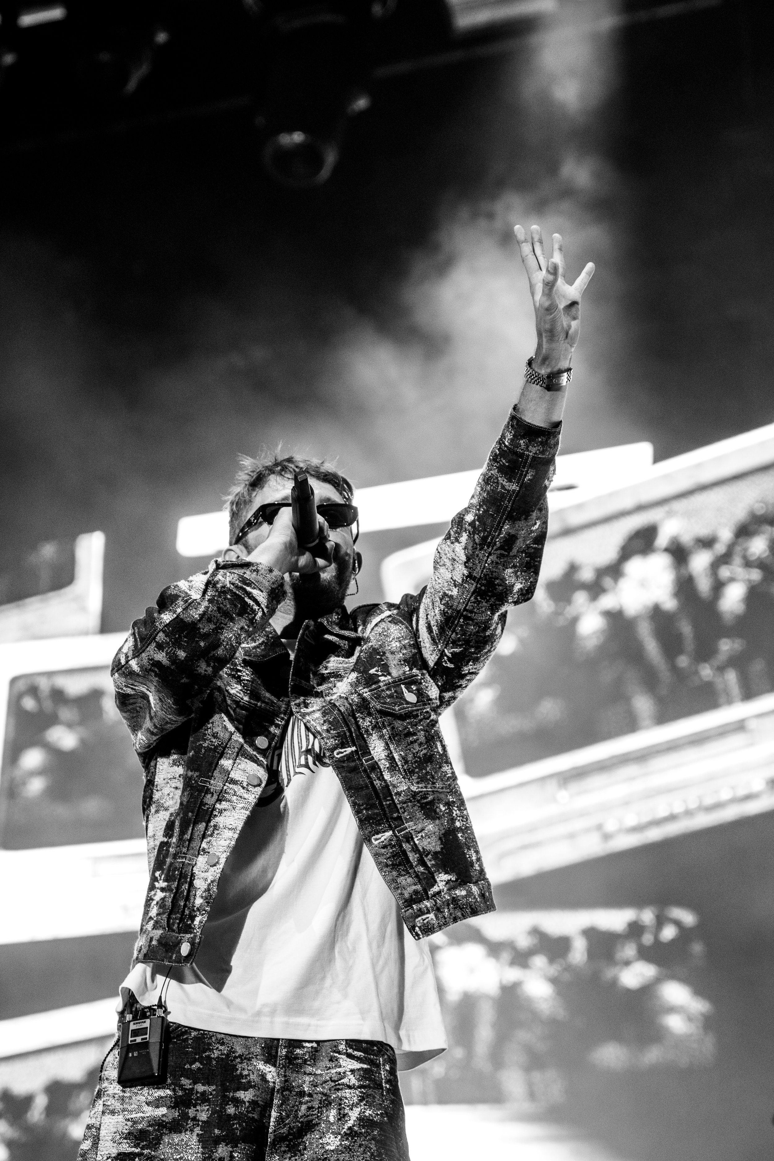 Un rappeur portant des lunettes de soleil, un jean déchiré et une veste à motifs, chante dans un micro avec un bras levé lors d'une performance sur scène, en noir et blanc.