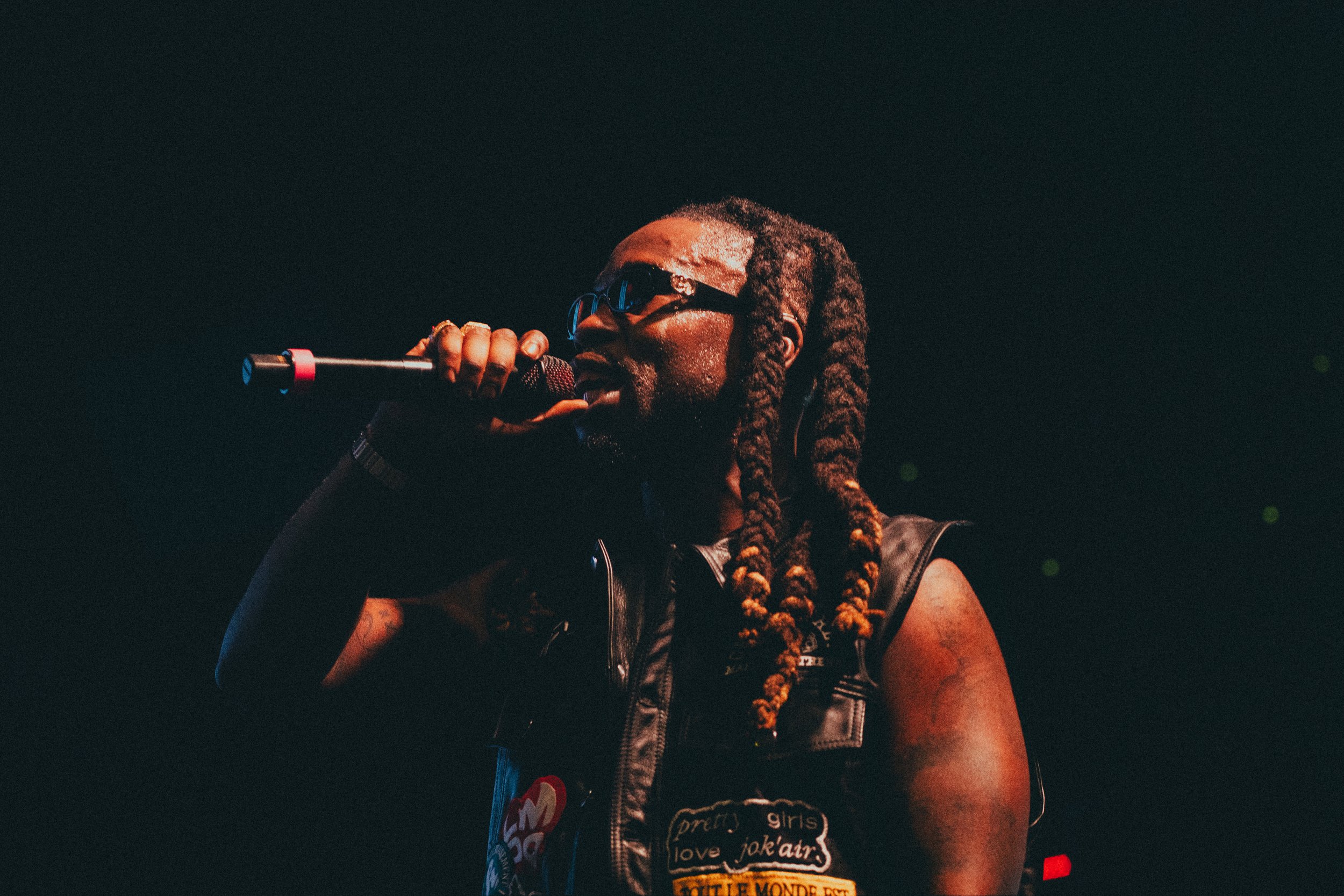 Un homme avec des dreadlocks portant des lunettes, tenant un microphone et chantant lors d'une performance, portant une veste en cuir avec des patches.