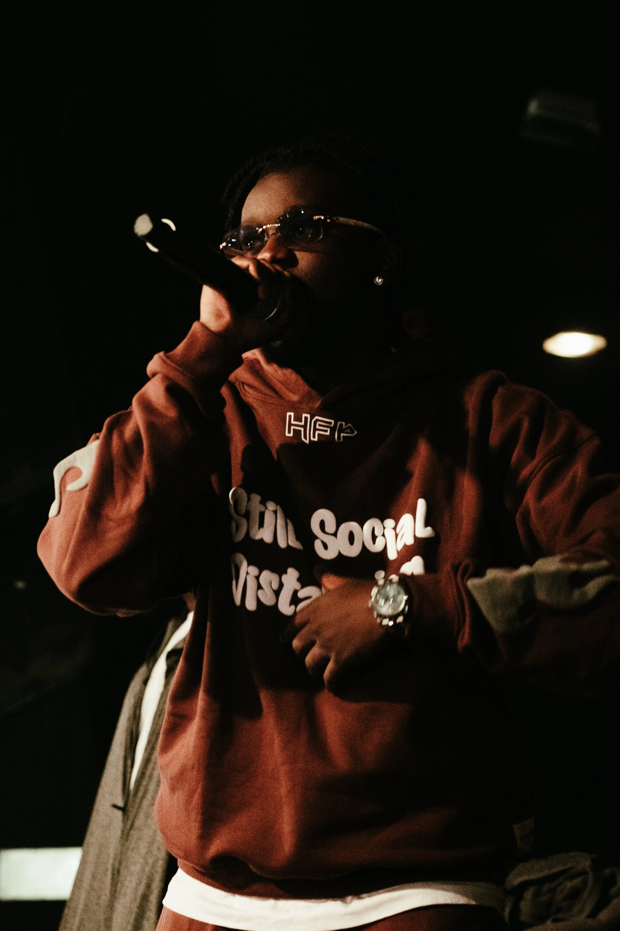 Une femme portant des lunettes de soleil et un sweat-shirt marron avec des inscriptions, tenant un microphone et apparemment en train de performer sur scène.