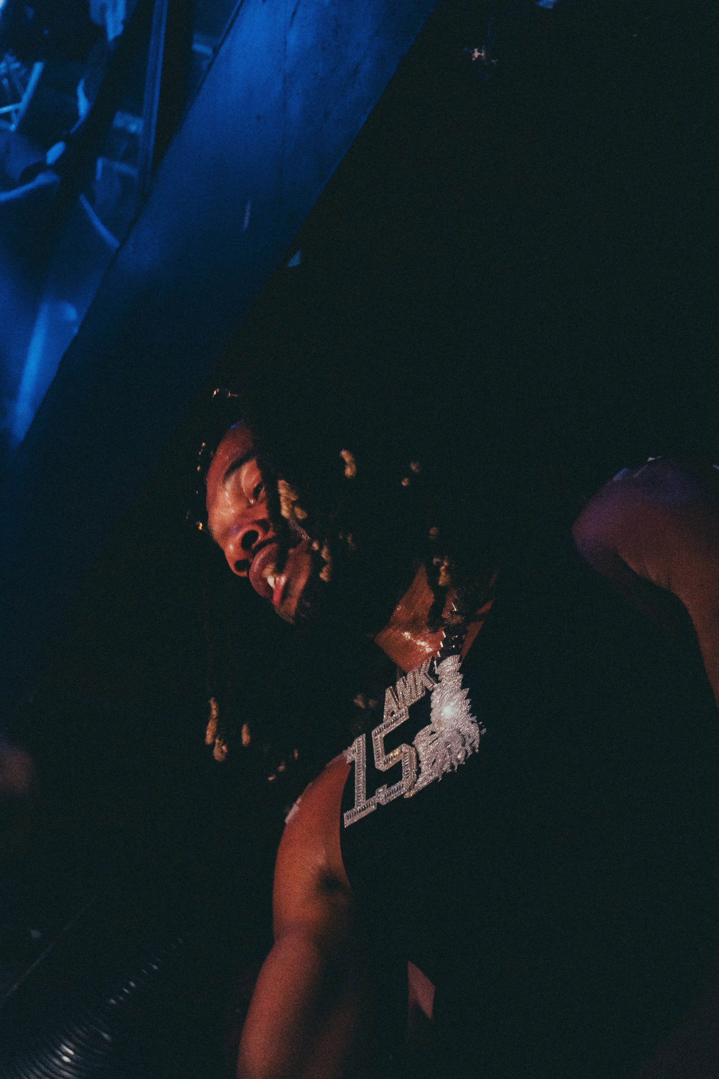 Un homme avec des dreadlocks portant un collier avec le texte '15' dans un environnement sombre, probablement un club ou une scène musicale.
