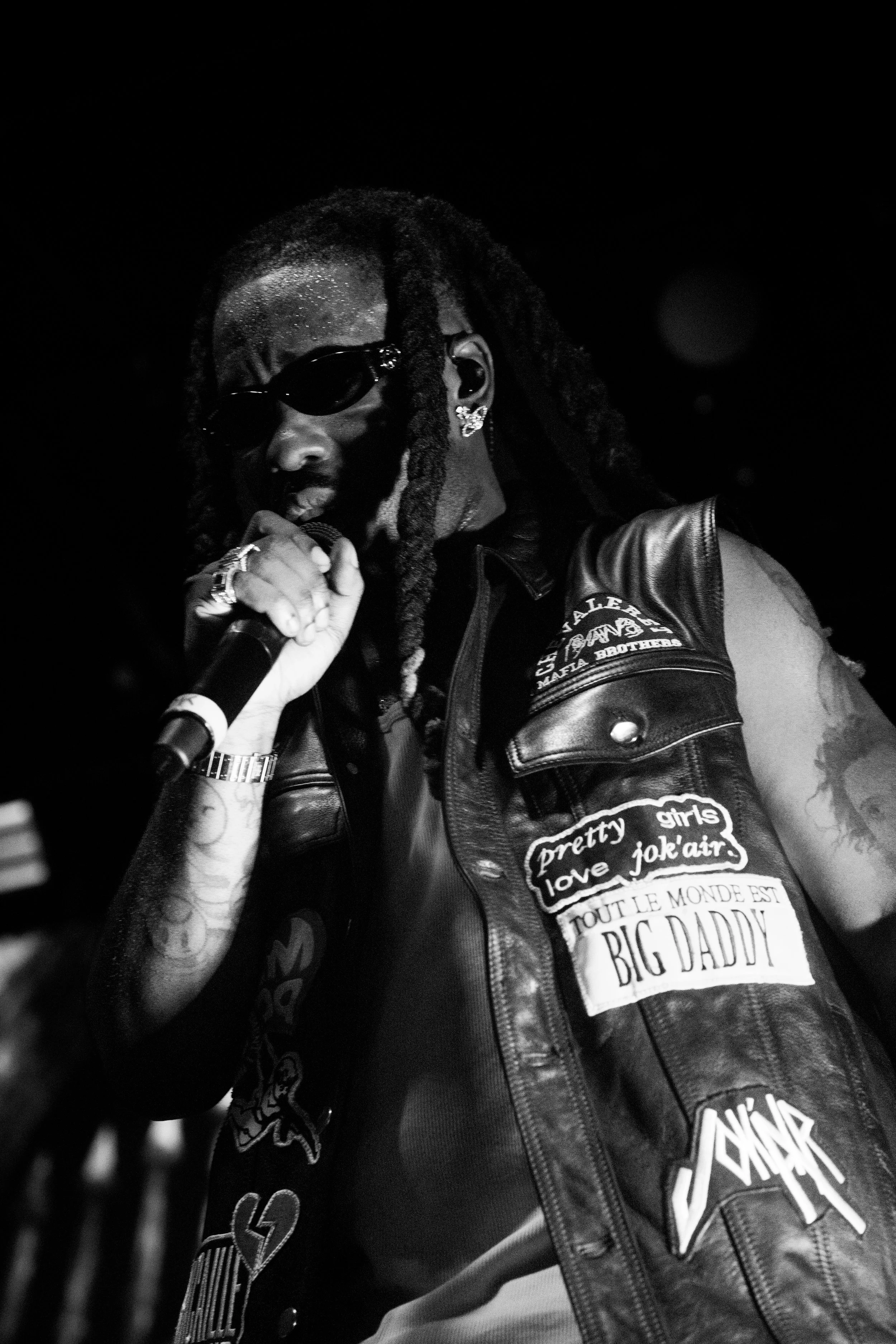 Un artiste de rap avec des dreadlocks portant des lunettes de soleil, un gilet en cuir orné de patchs, tenant un microphone lors d'une performance.