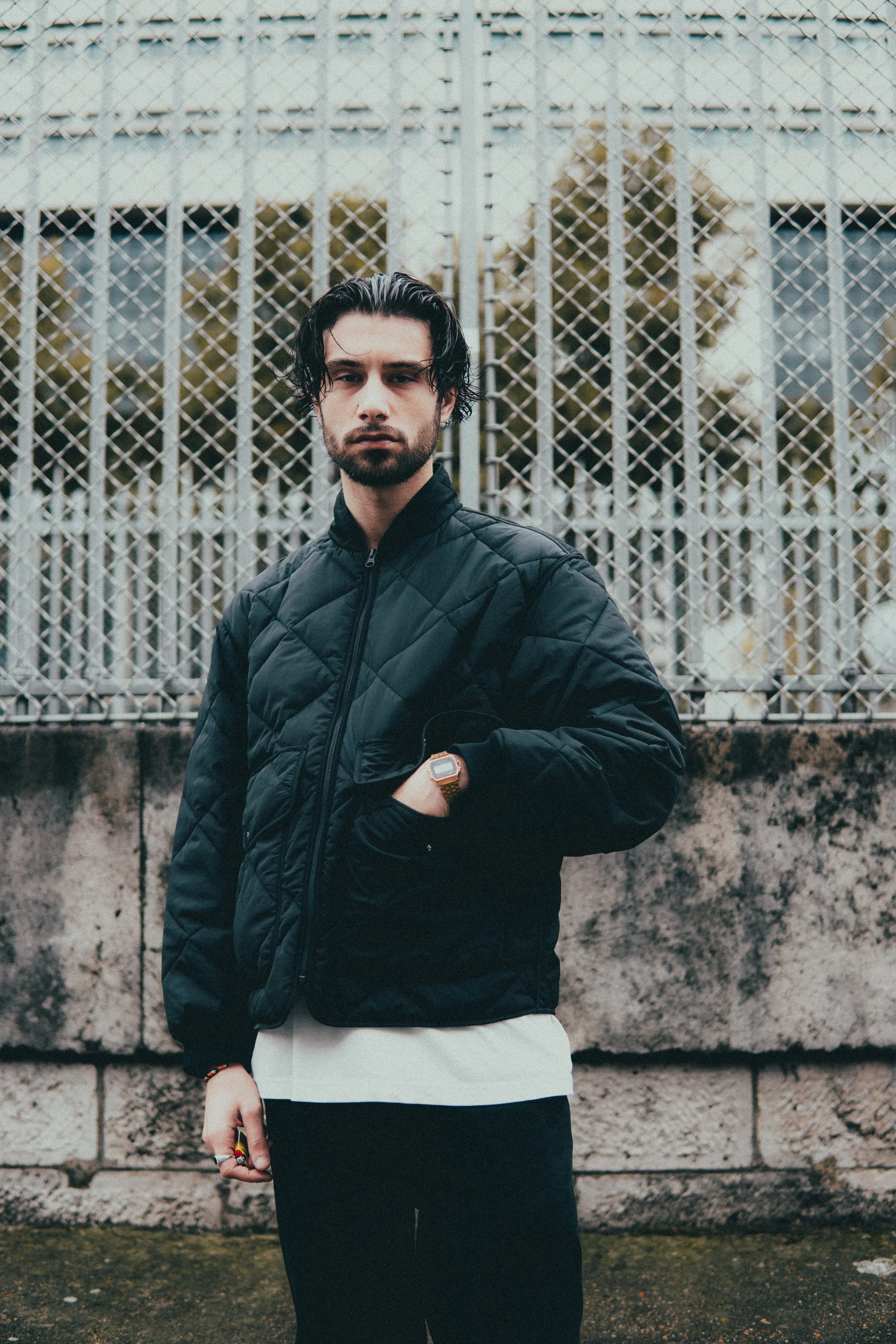 Un jeune homme en manteau noir, debout devant une clôture en métal, avec un mur en pierre en arrière-plan.