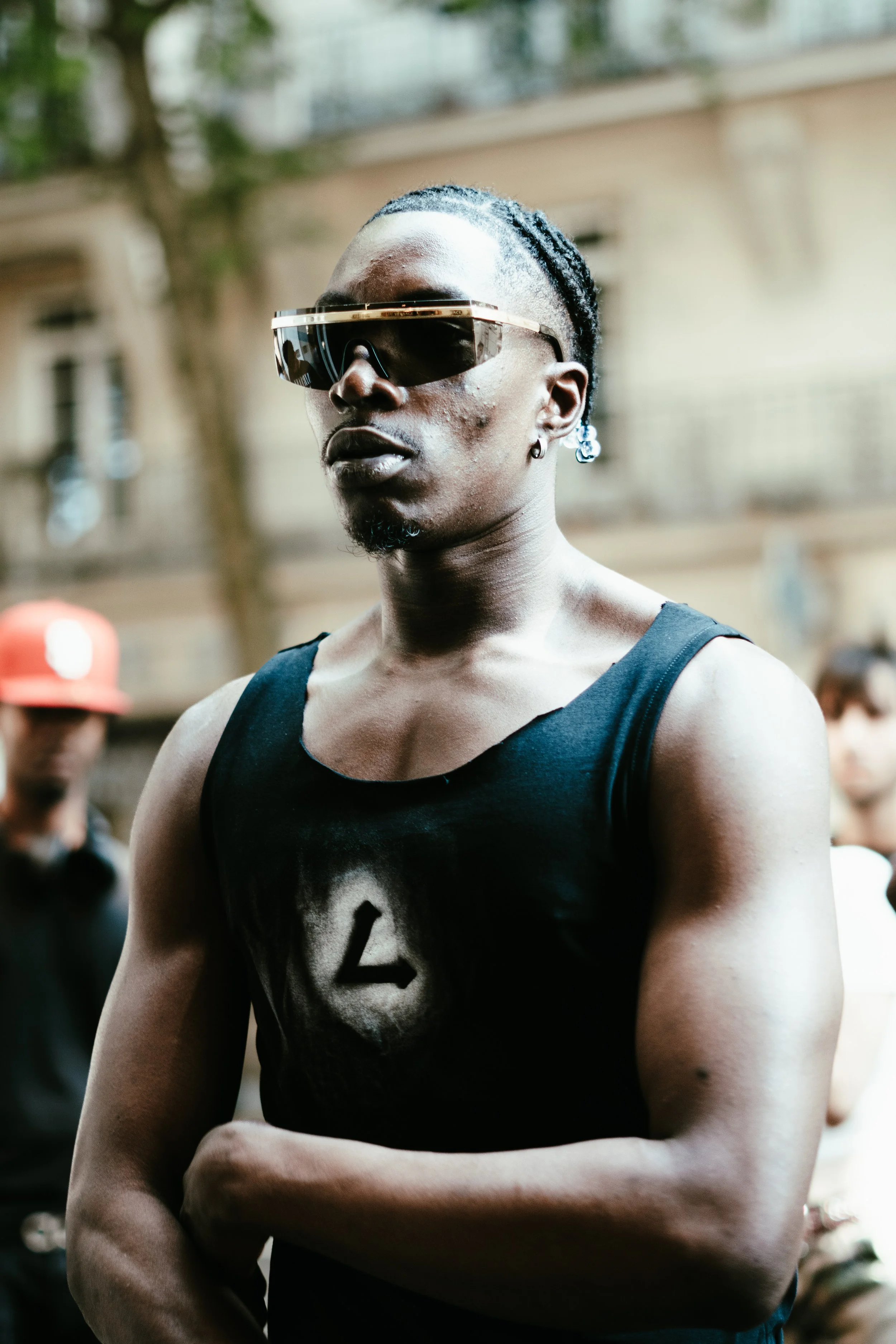 Un jeune homme portant des lunettes de soleil foncées et un t-shirt noir avec un symbole au centre, étant dans un espace extérieur avec d'autres personnes en arrière-plan.