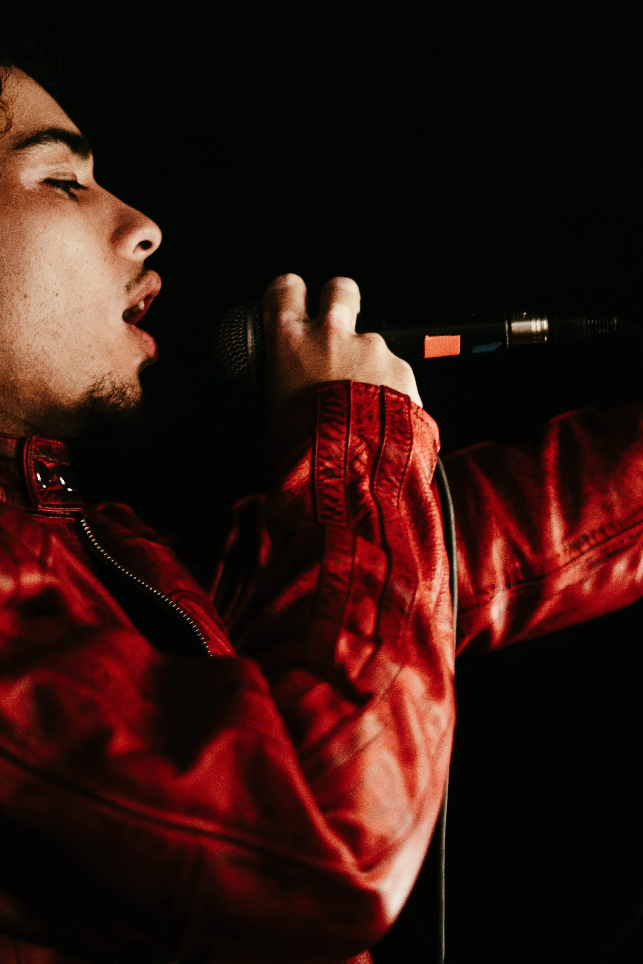Un jeune homme chantant ou parlant dans un microphone, habillé d'une veste en cuir rouge, sur un fond noir.