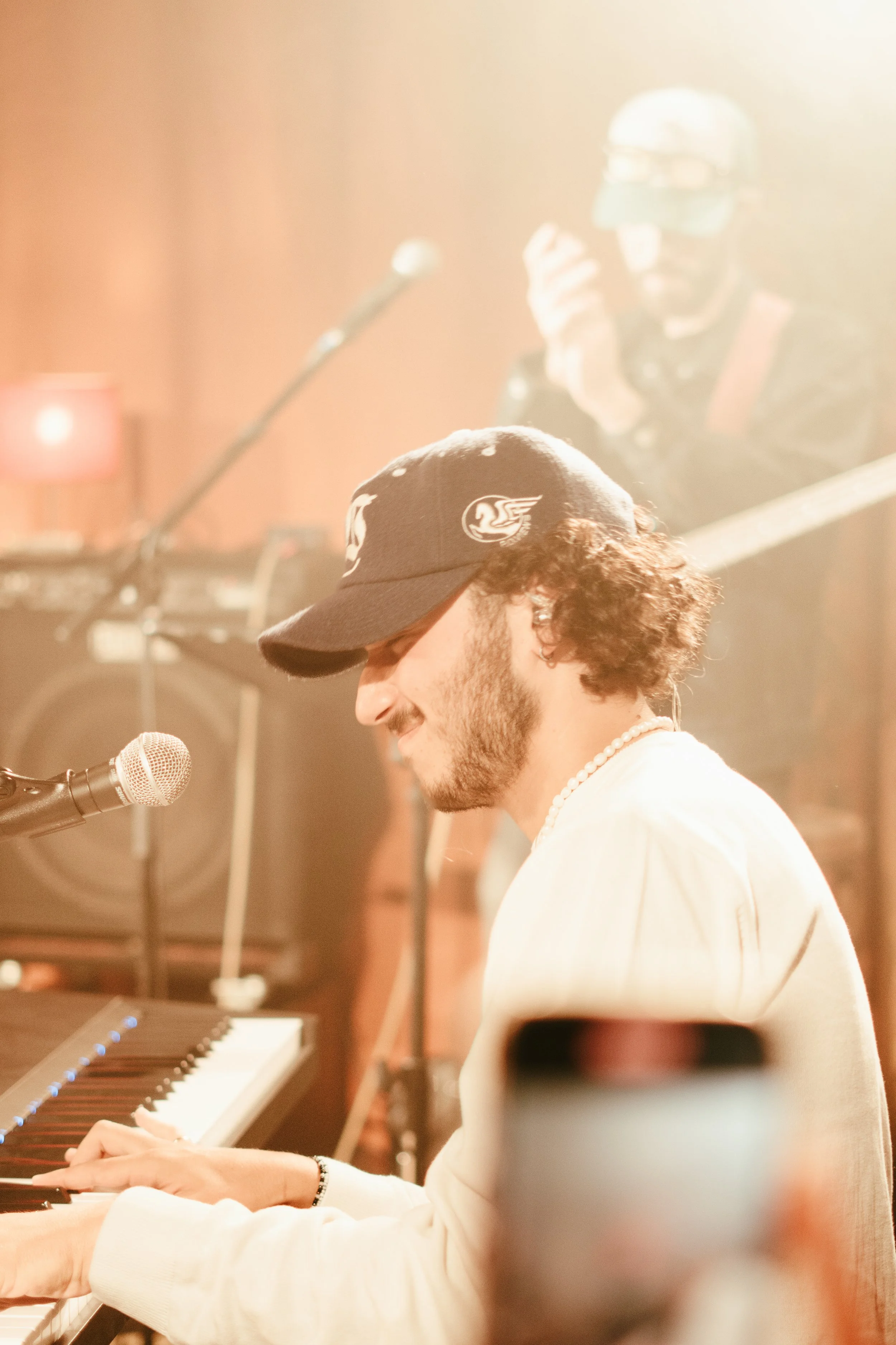 Un homme joue du piano lors d'une performance, portant une casquette noire, des cheveux bouclés, et une chaîne de perles, avec un musicien en arrière-plan portant un casque et une guitare électrique.